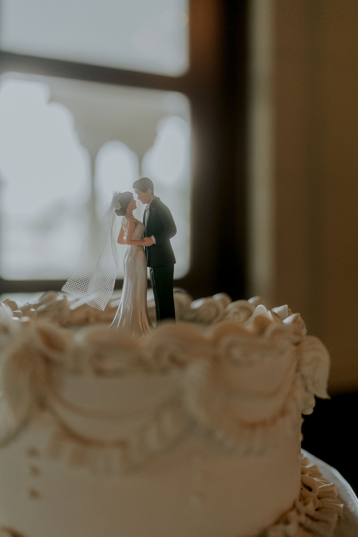 Classic white wedding cake topped with bride and groom figurines in soft window light.