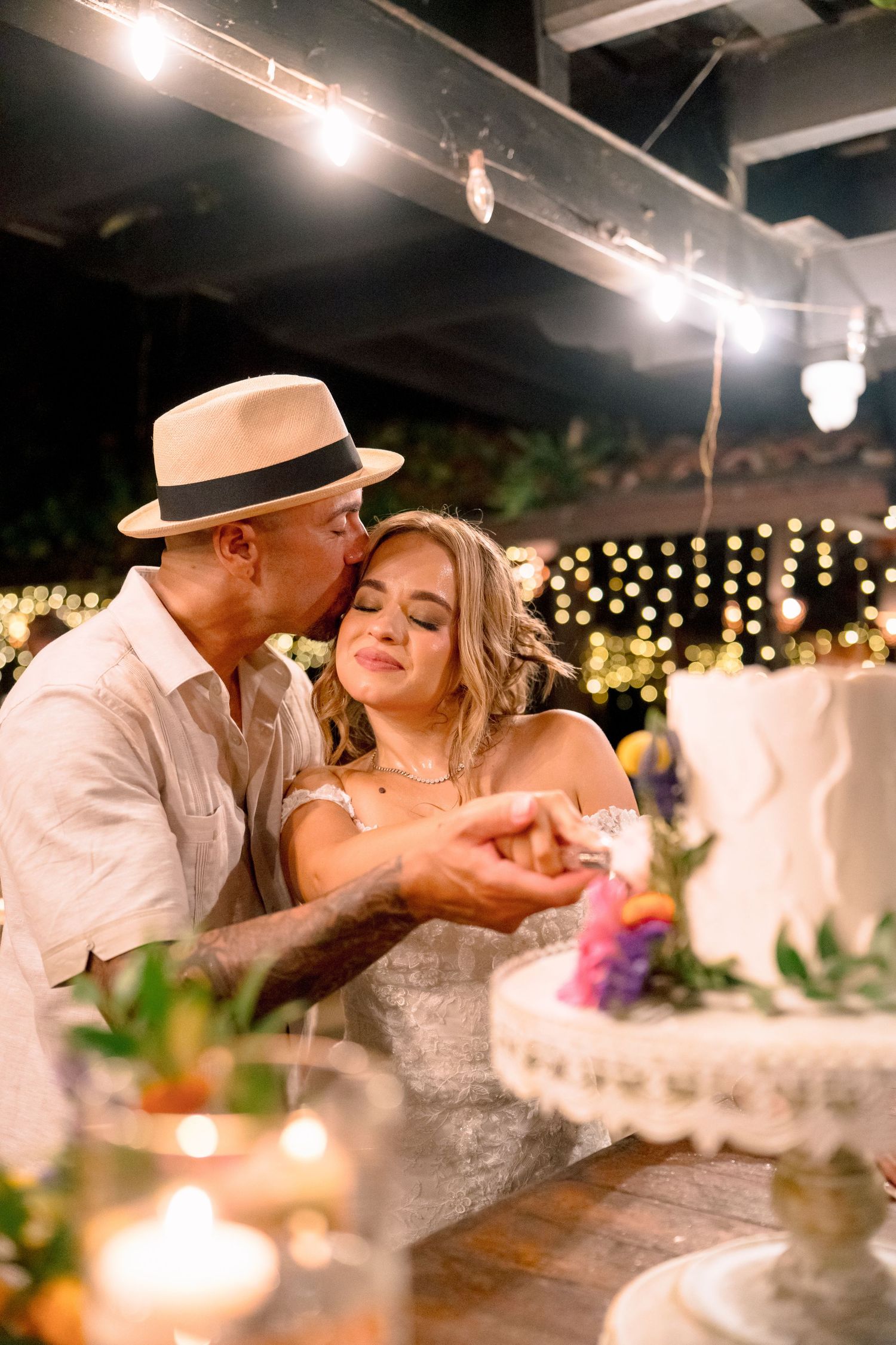 Groom in panama hat kissing bride on cheek while cutting white wedding cake under string lights.