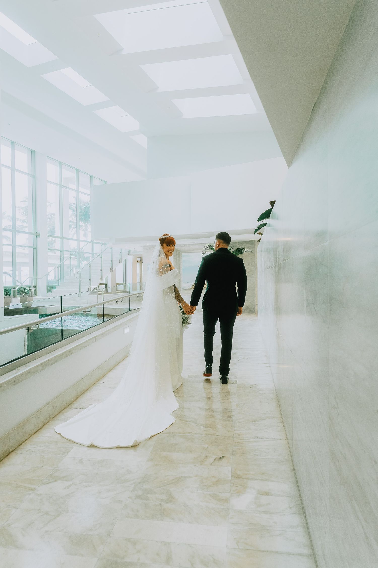 Bride in white gown and groom in black suit hold hands walking through elegant marble wedding venue.