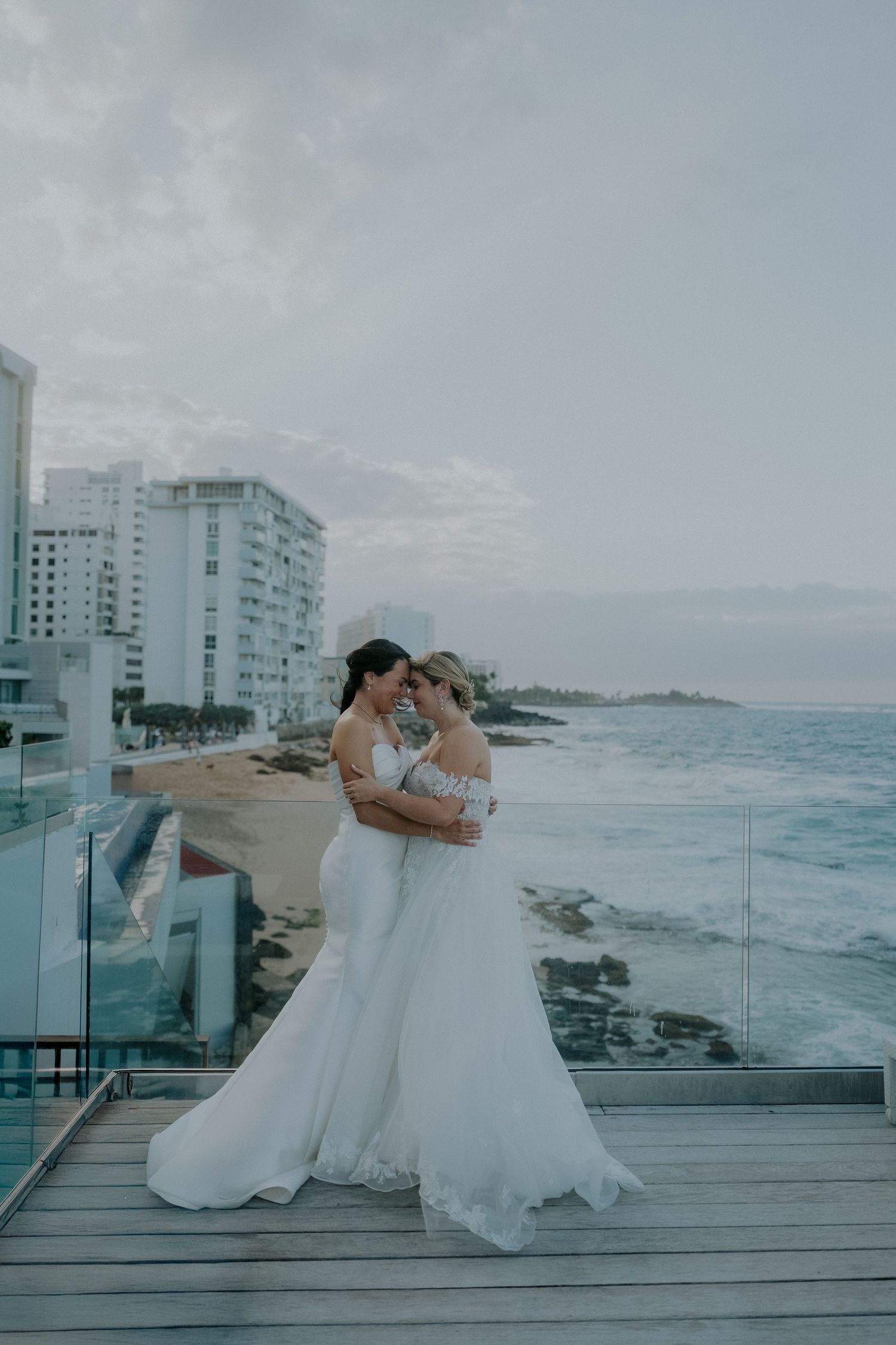 Two brides in white gowns embrace on a seaside balcony with ocean waves and resort buildings in the background.