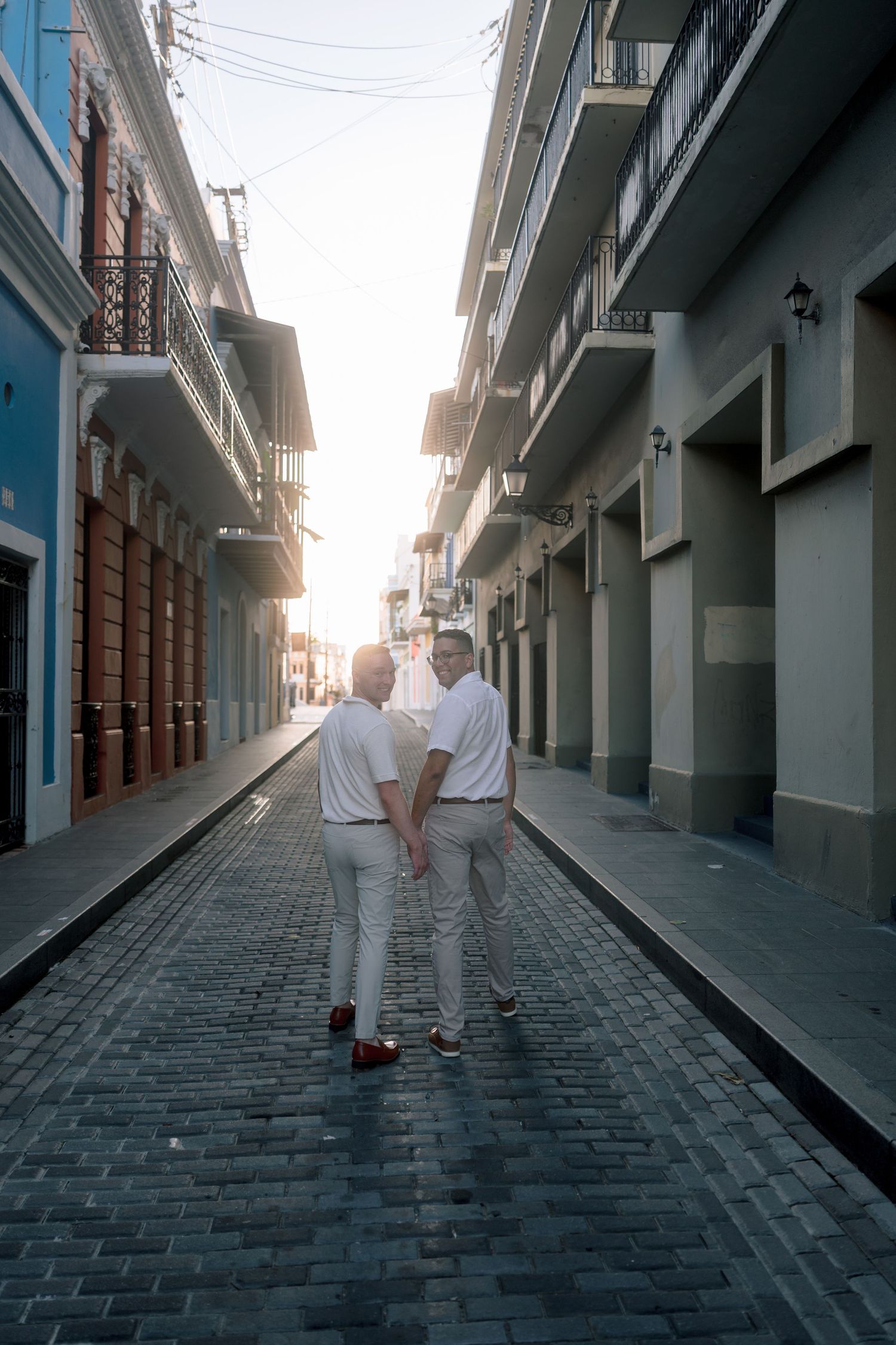 Two men holding hands walk down a sunlit cobblestone street in Old San Juan, Puerto Rico, wearing matching white outfits.