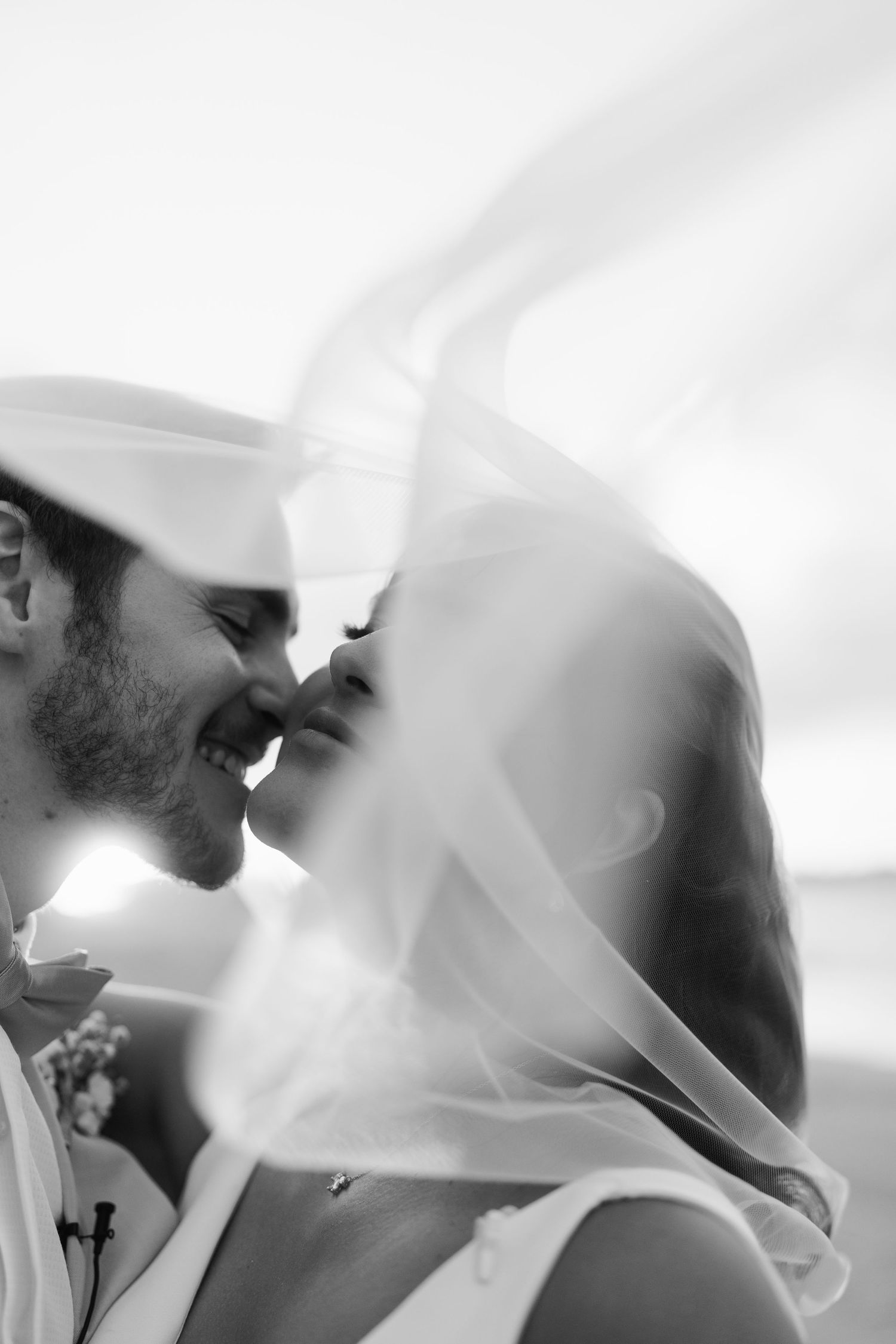 Black and white romantic photo of a smiling couple nearly kissing under a billowing bridal veil at the beach.
