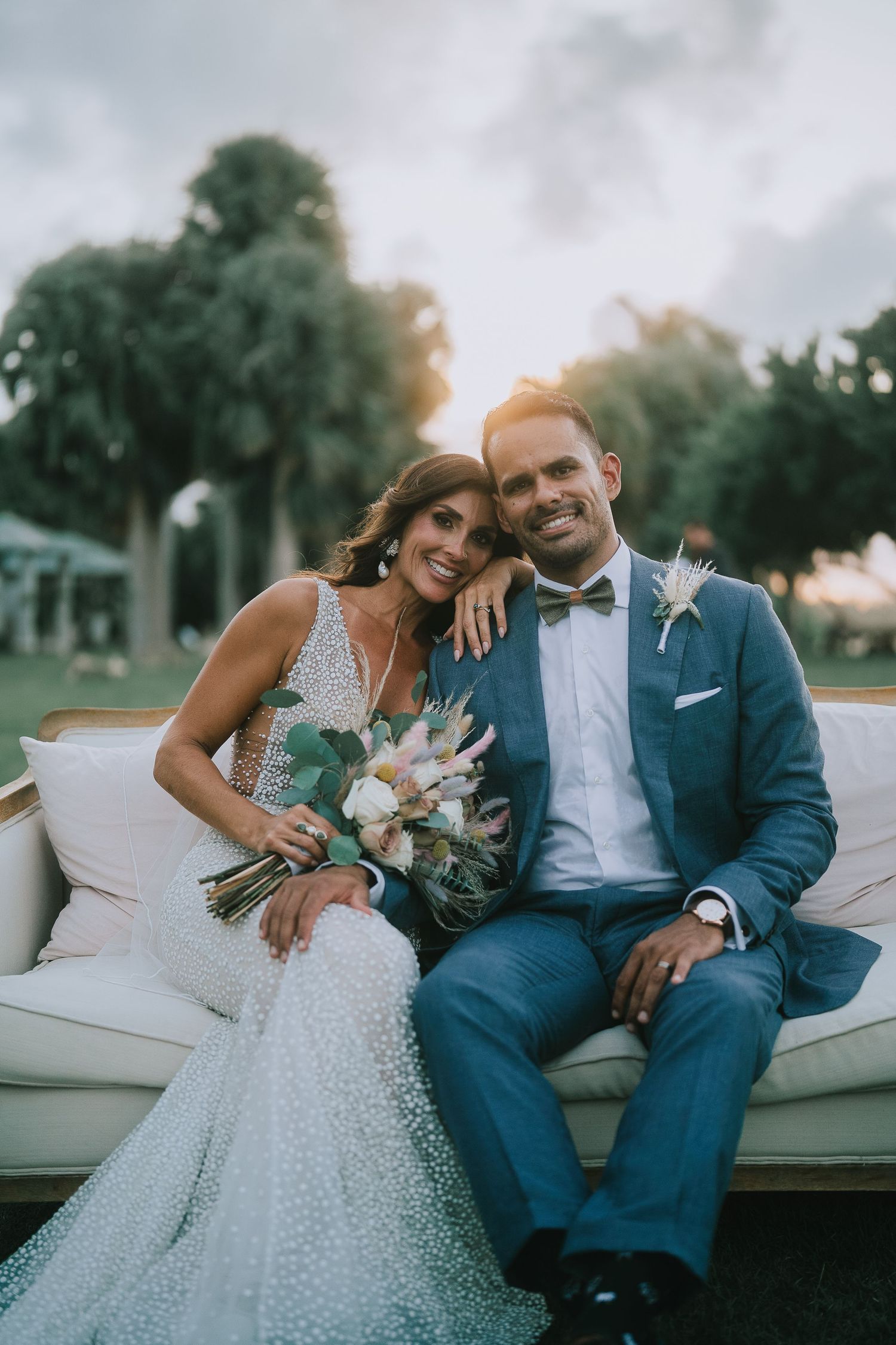 Bride and groom sitting on white sofa at sunset with tropical background.