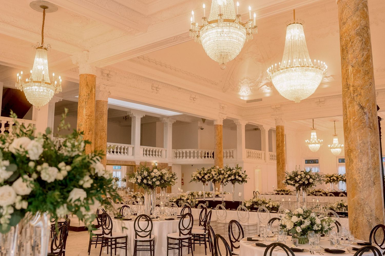 Elegant wedding reception hall with crystal chandeliers, marble columns, and floral centerpieces on long decorated tables.