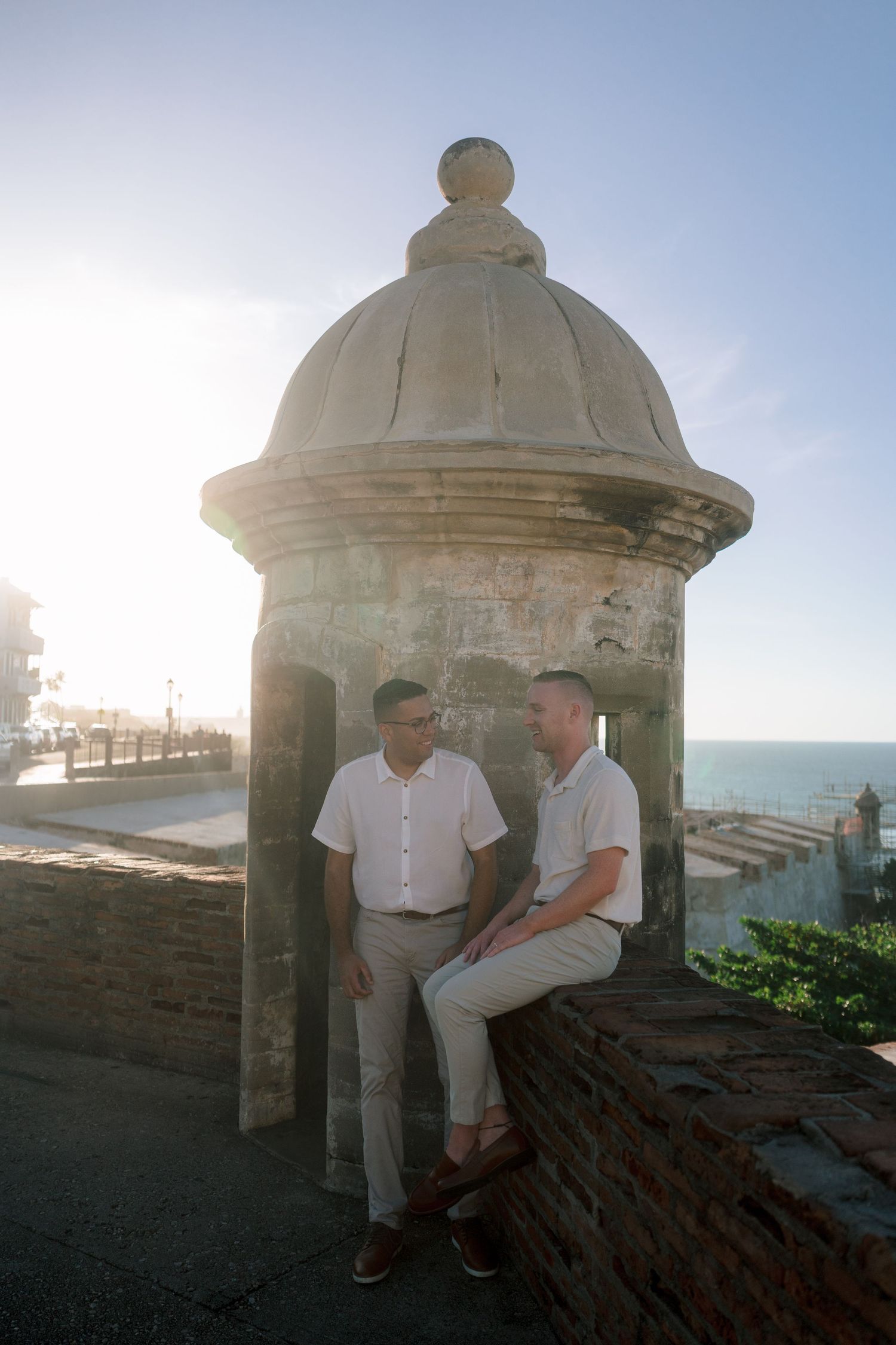 Two men in white shirts sit by a historic stone sentry box in Old San Juan, Puerto Rico, with ocean views.