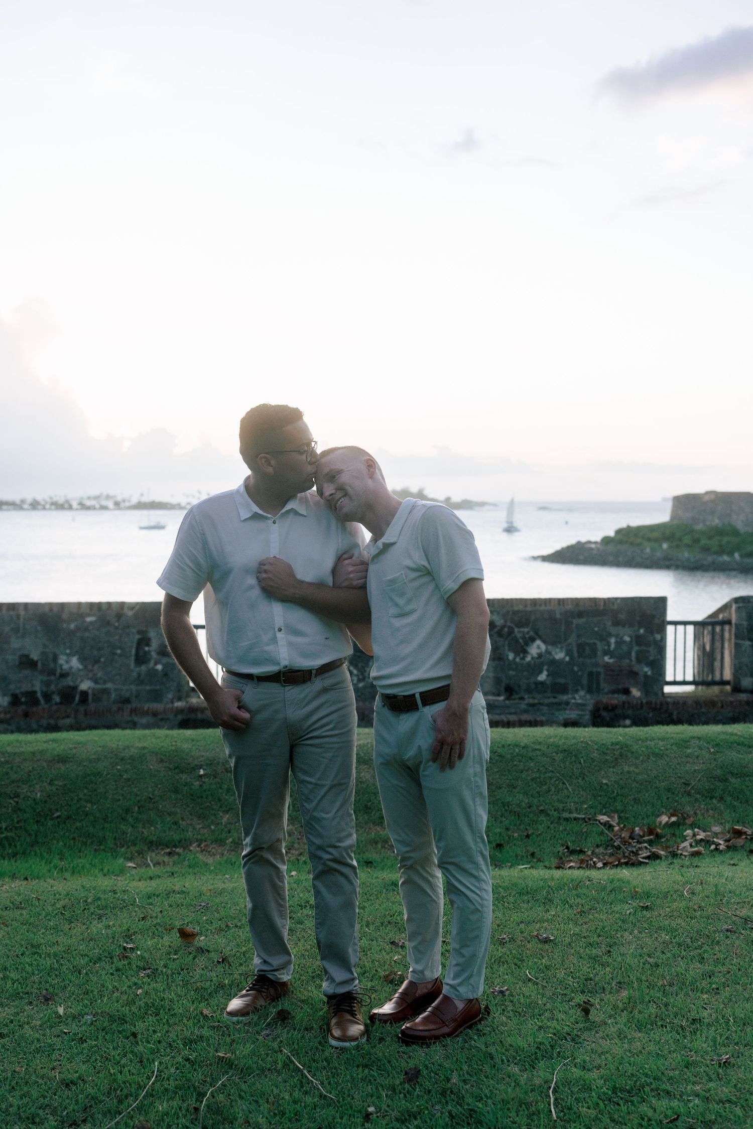 Two men share a tender moment on a grassy area with a scenic waterfront and historic fort in the background.
