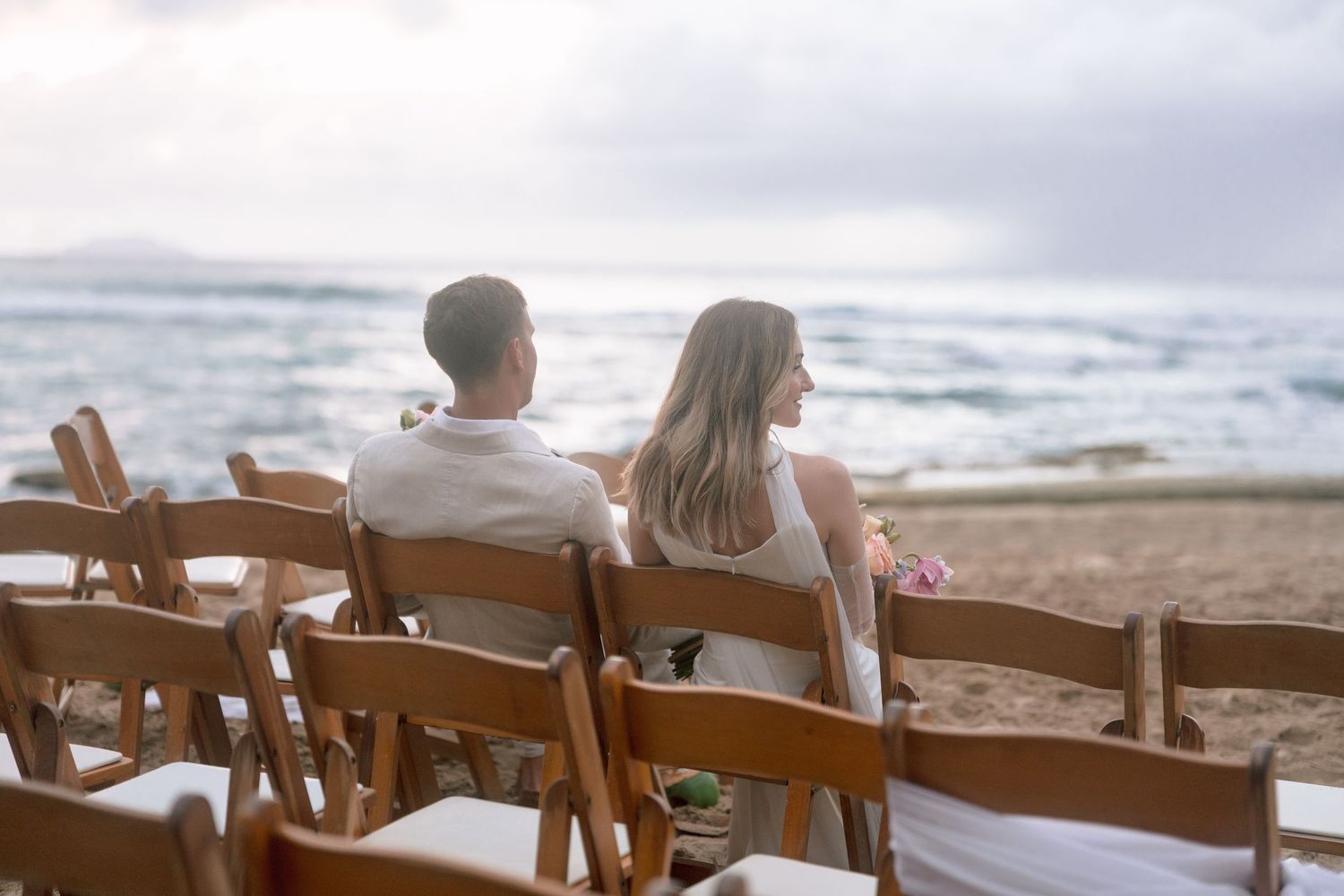 Bride and groom sit together on wooden chairs facing the ocean at a beach wedding ceremony.