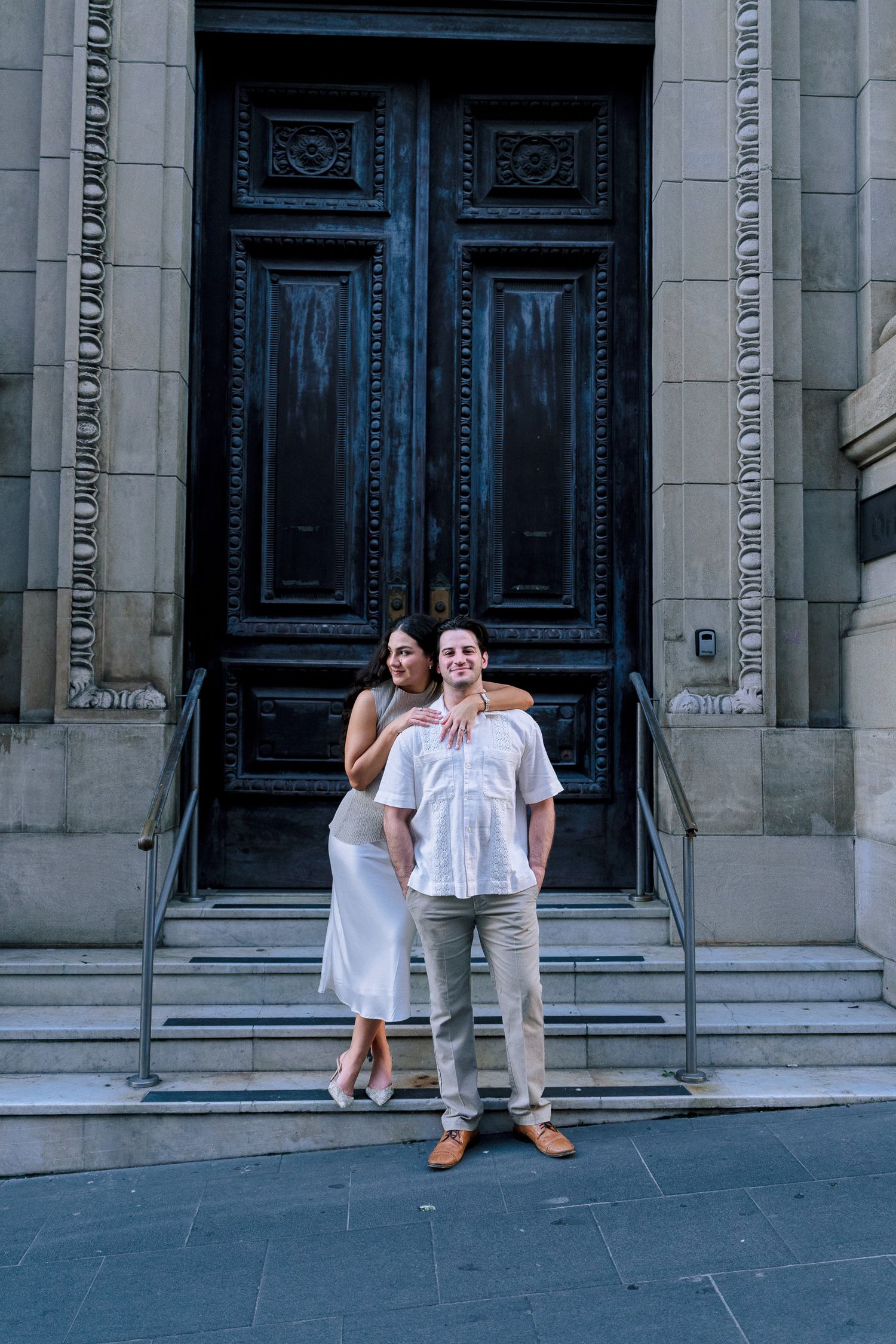 Couple in white outfits posing on steps before ornate dark wooden doors of a historic building.