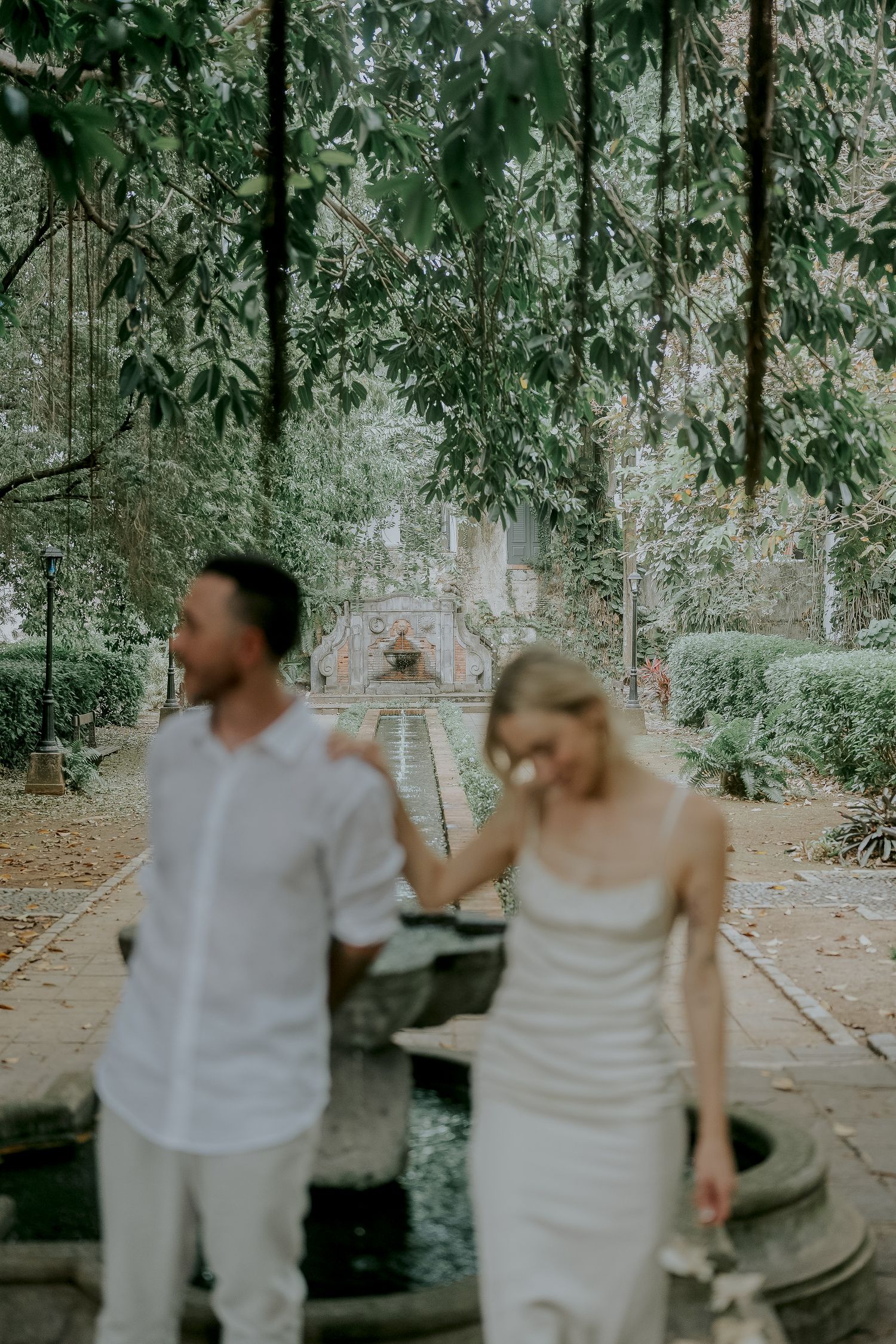 Couple holding hands walking through a lush garden with a fountain and greenery backdrop.