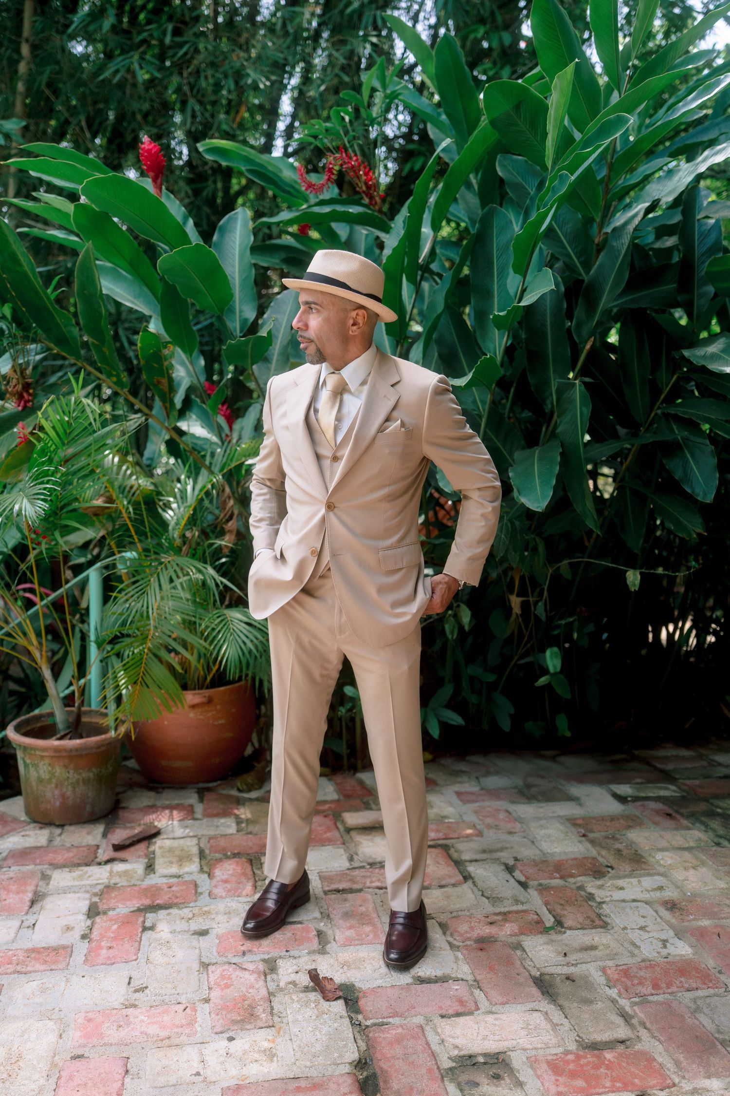 A stylish man in a beige suit and straw hat poses confidently in a lush tropical garden setting.