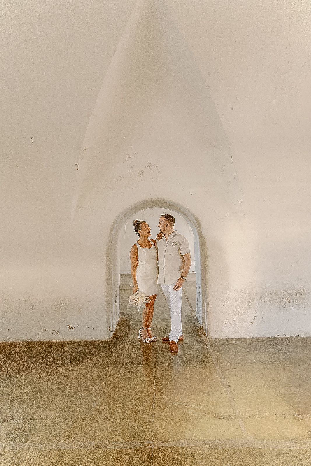Couple in white attire sharing a romantic moment under an arch in a sunlit corridor.