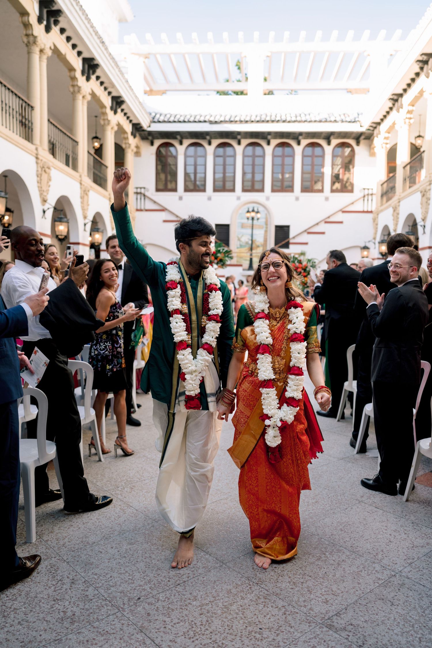 Couple in traditional Indian wedding attire walks joyfully down aisle in courtyard venue.