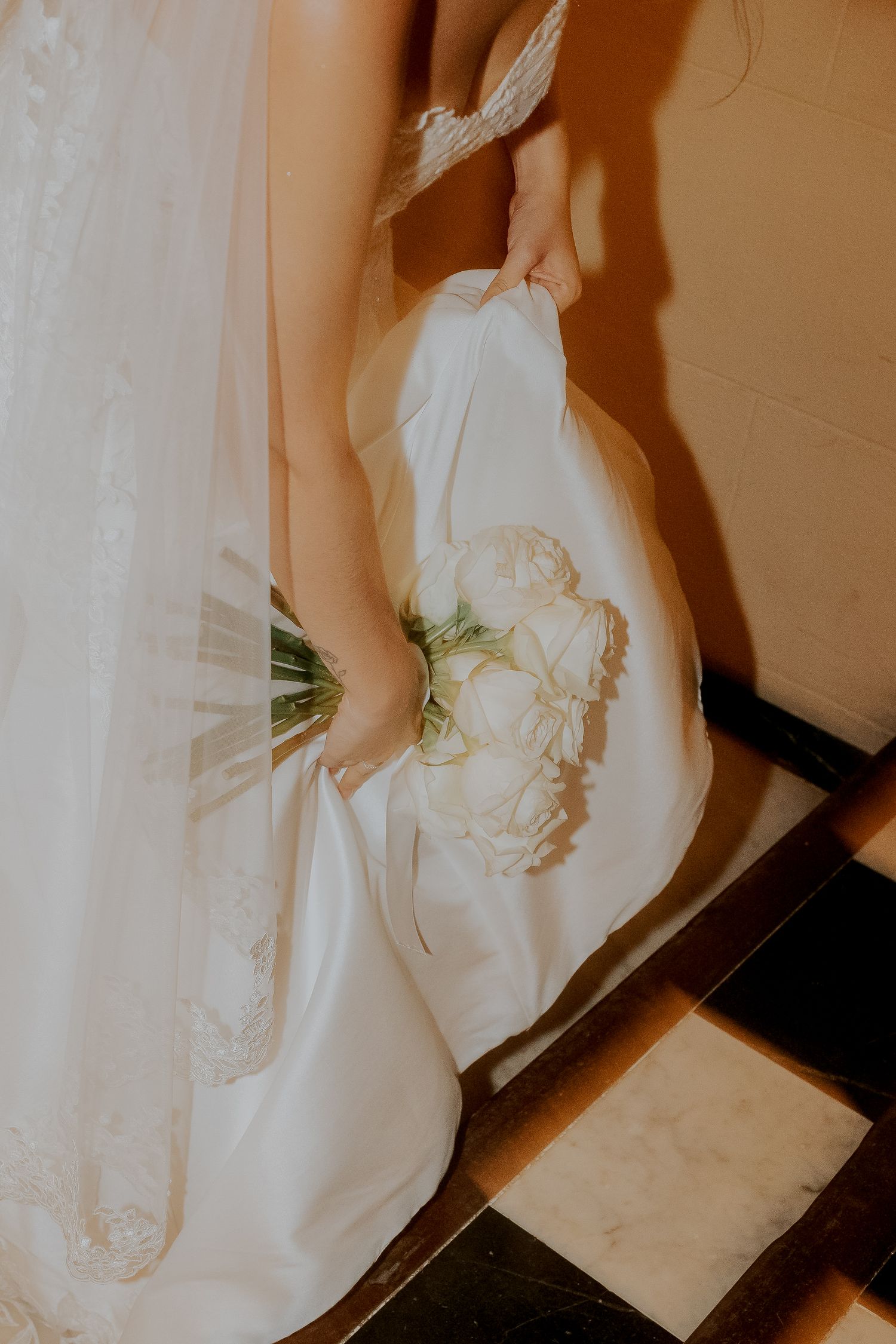 Close-up of bride holding white rose bouquet and gathering lace wedding gown train indoors.