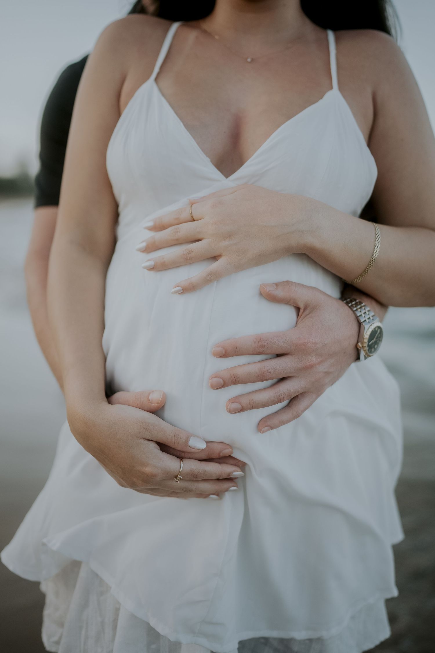Pregnant woman in white dress with partner's hands cradling baby bump during maternity photoshoot.