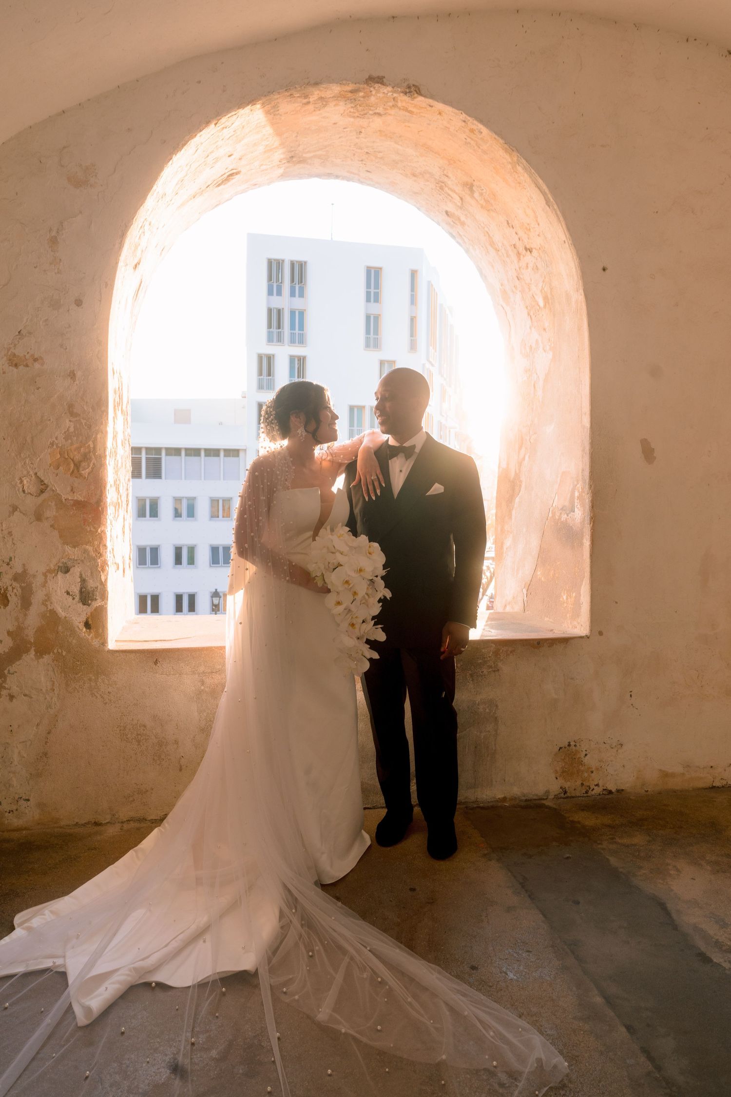 Bride and groom pose in a sunlit arched window alcove, bride holding white bouquet with flowing train.