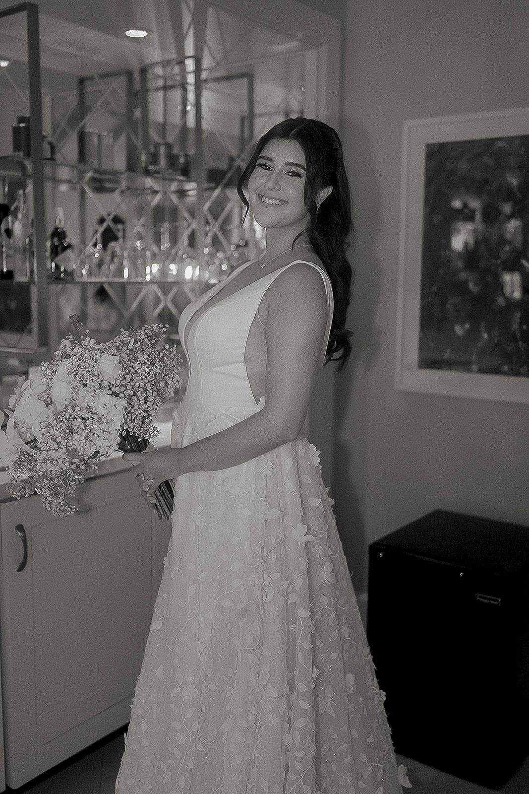 Smiling bride in floral wedding gown holding bouquet in black and white photo.