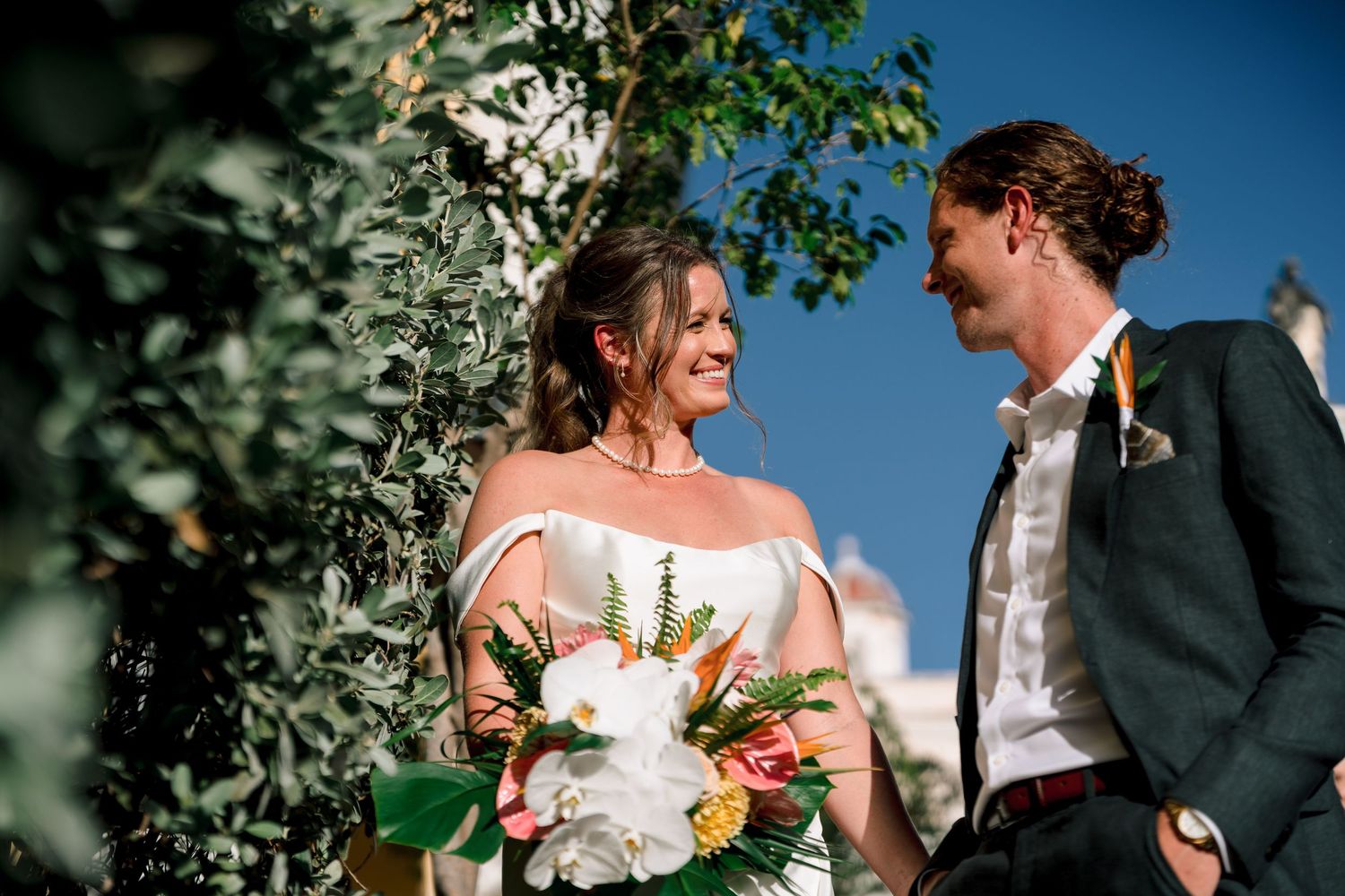 Bride and groom share a romantic moment under lush greenery, bride holding a tropical bouquet on their wedding day.