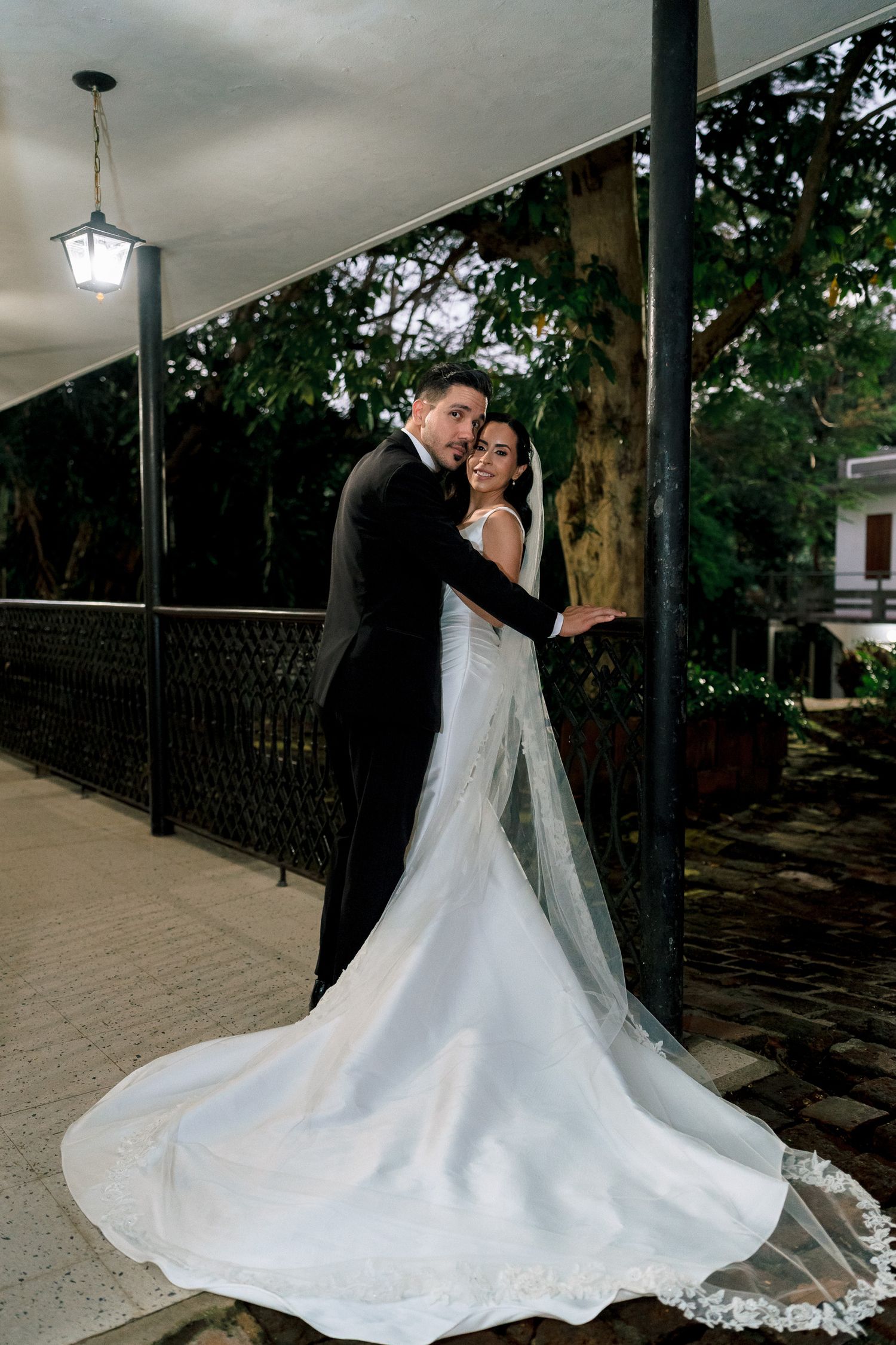 A bride in a flowing white gown with a long train poses with a groom in a black tuxedo on an outdoor walkway.
