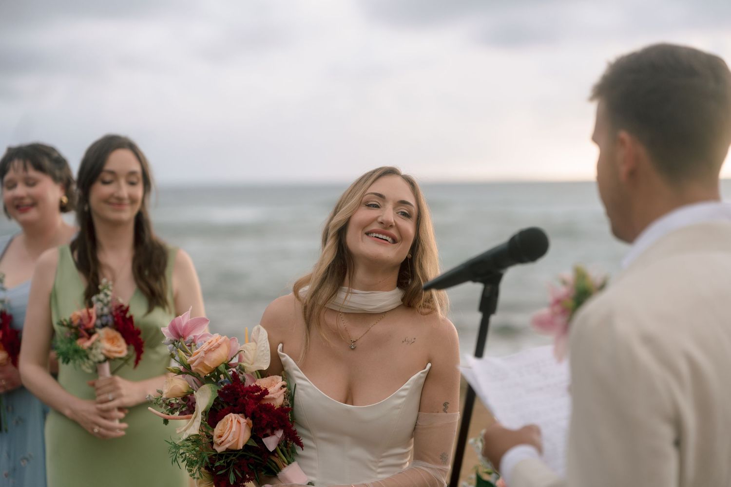 Bride smiling joyfully at beach wedding ceremony with bridesmaids in green dresses holding colorful bouquets by the ocean.