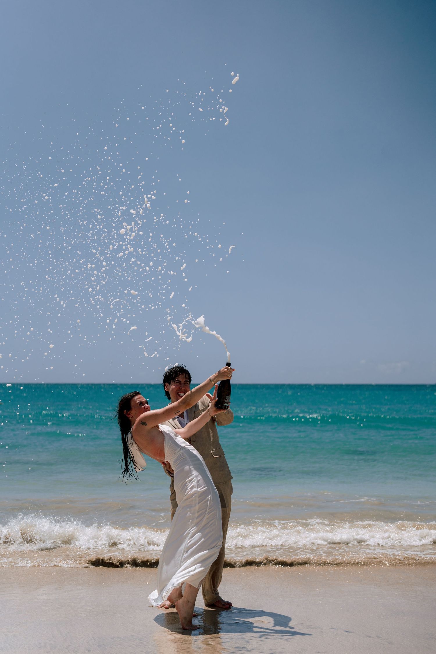 Joyful newlyweds celebrate on a sunny beach, popping champagne bottle with ocean waves behind them.