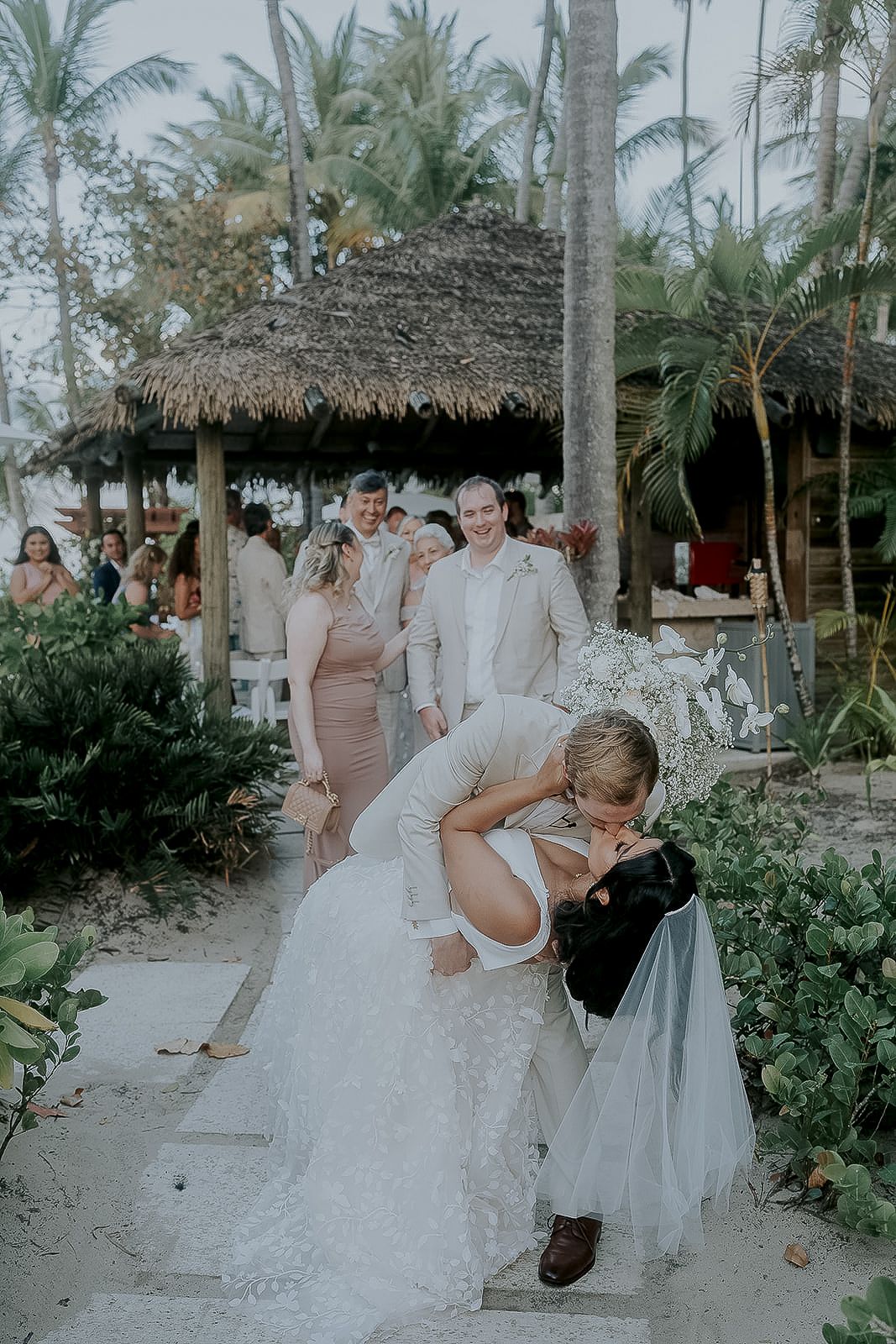 Groom dips bride for romantic kiss at tropical beach wedding ceremony with palm trees and guests.