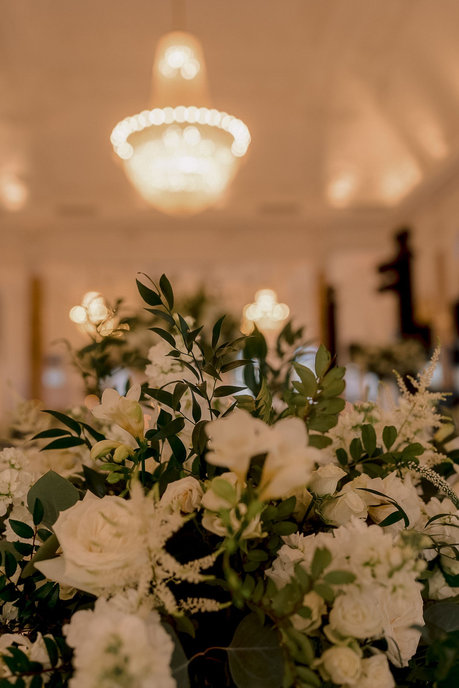 Elegant white floral centerpiece with roses and greenery under a glowing chandelier in a wedding reception hall.