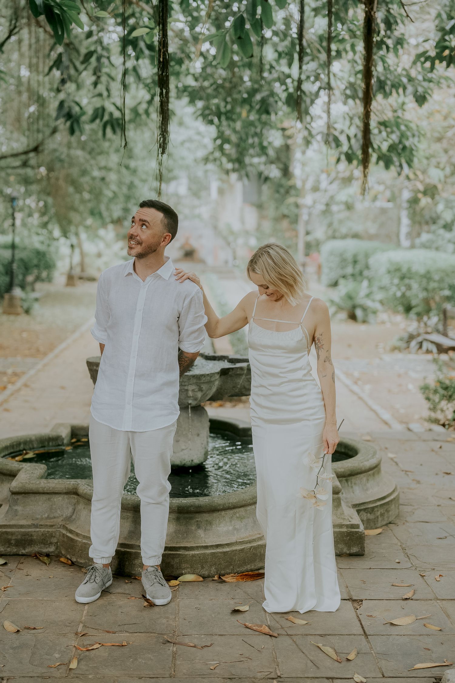 Couple in white outfits posing by a stone fountain in a lush garden setting for an elegant outdoor photoshoot.