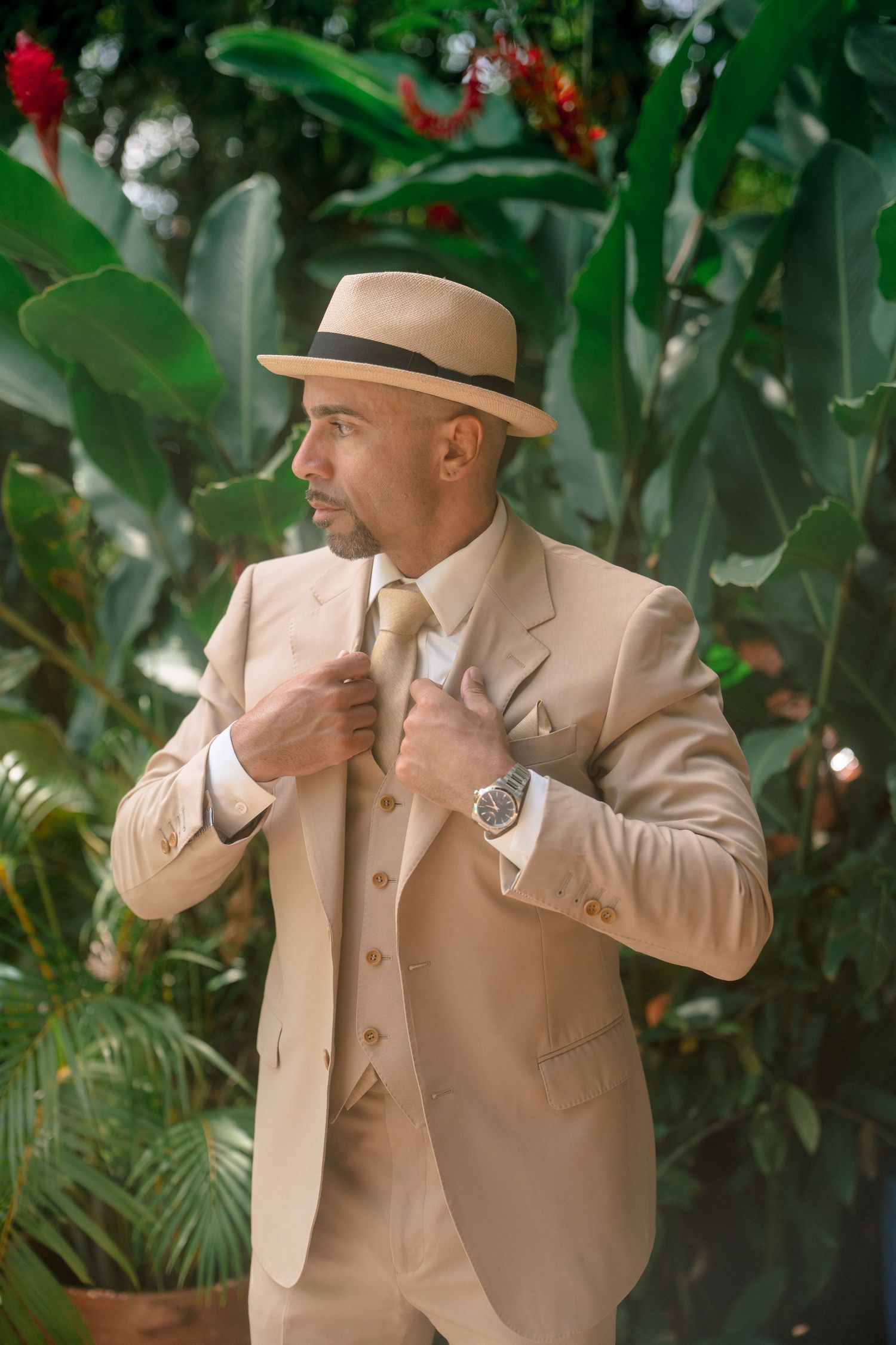 Stylish man in beige three-piece suit and panama hat adjusting tie in tropical garden setting.