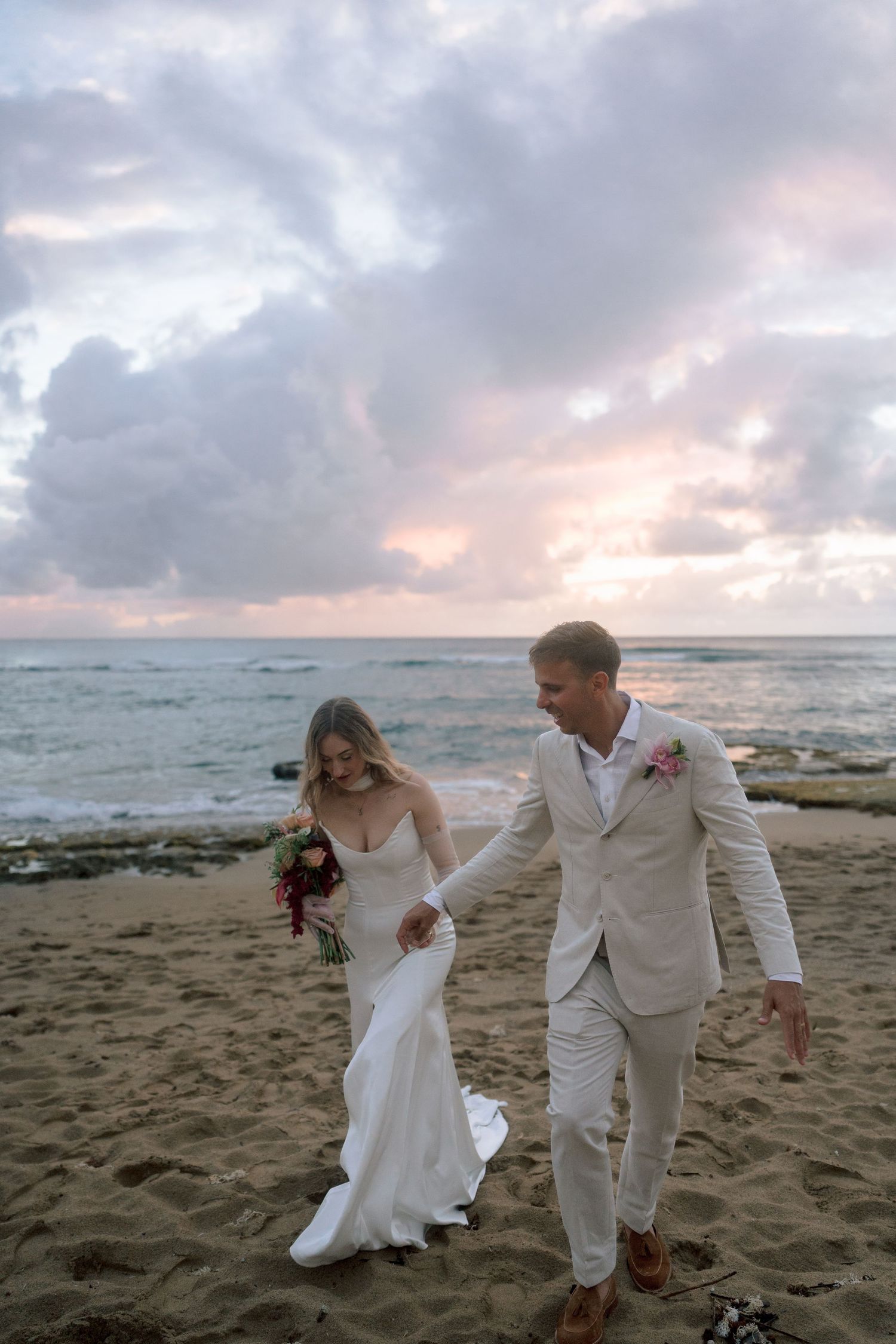 Laughing newlyweds holding hands strolling along beach at sunset, bride in fitted white gown with train.