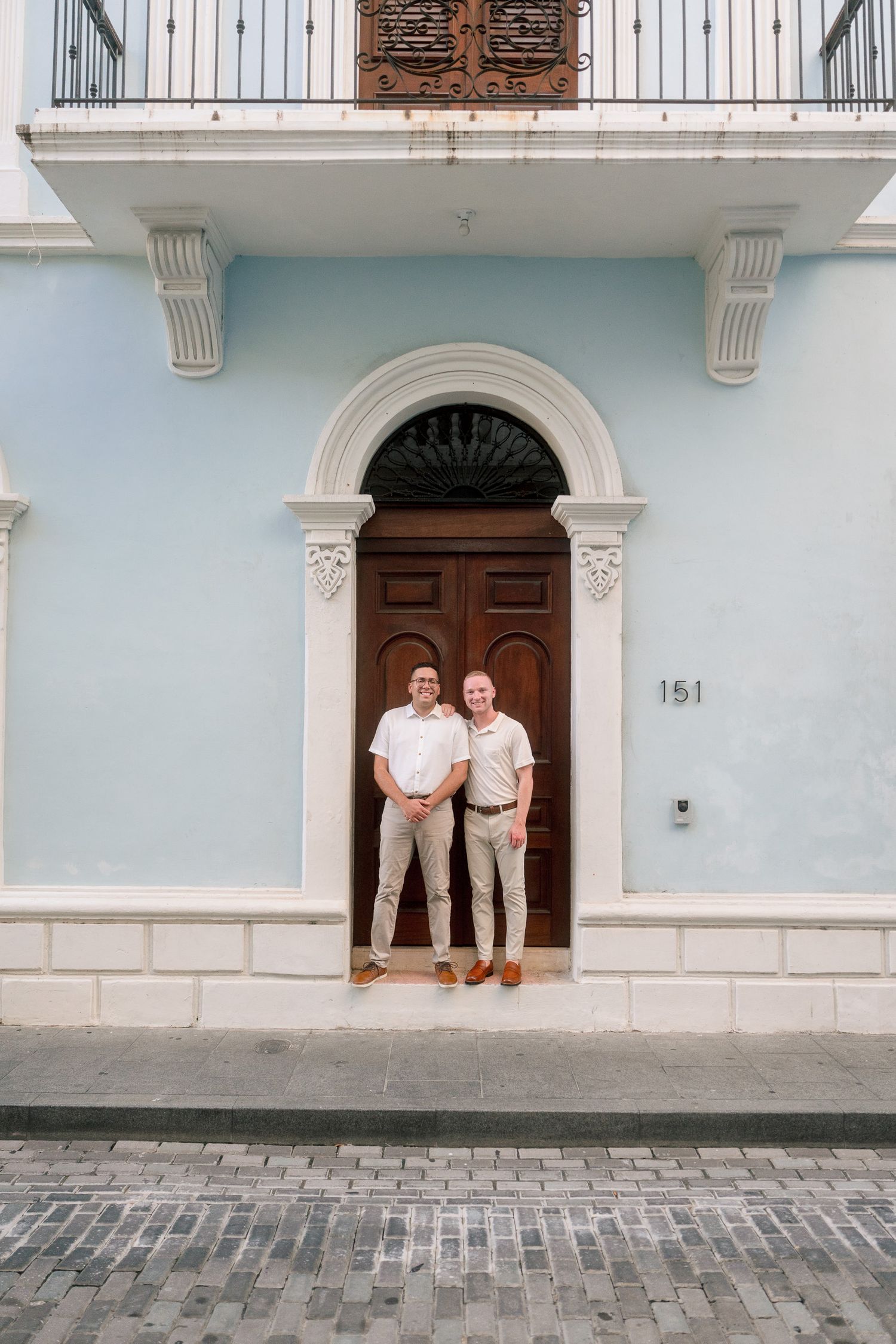 Two men smiling in front of a blue colonial building with arched wooden door at number 151.