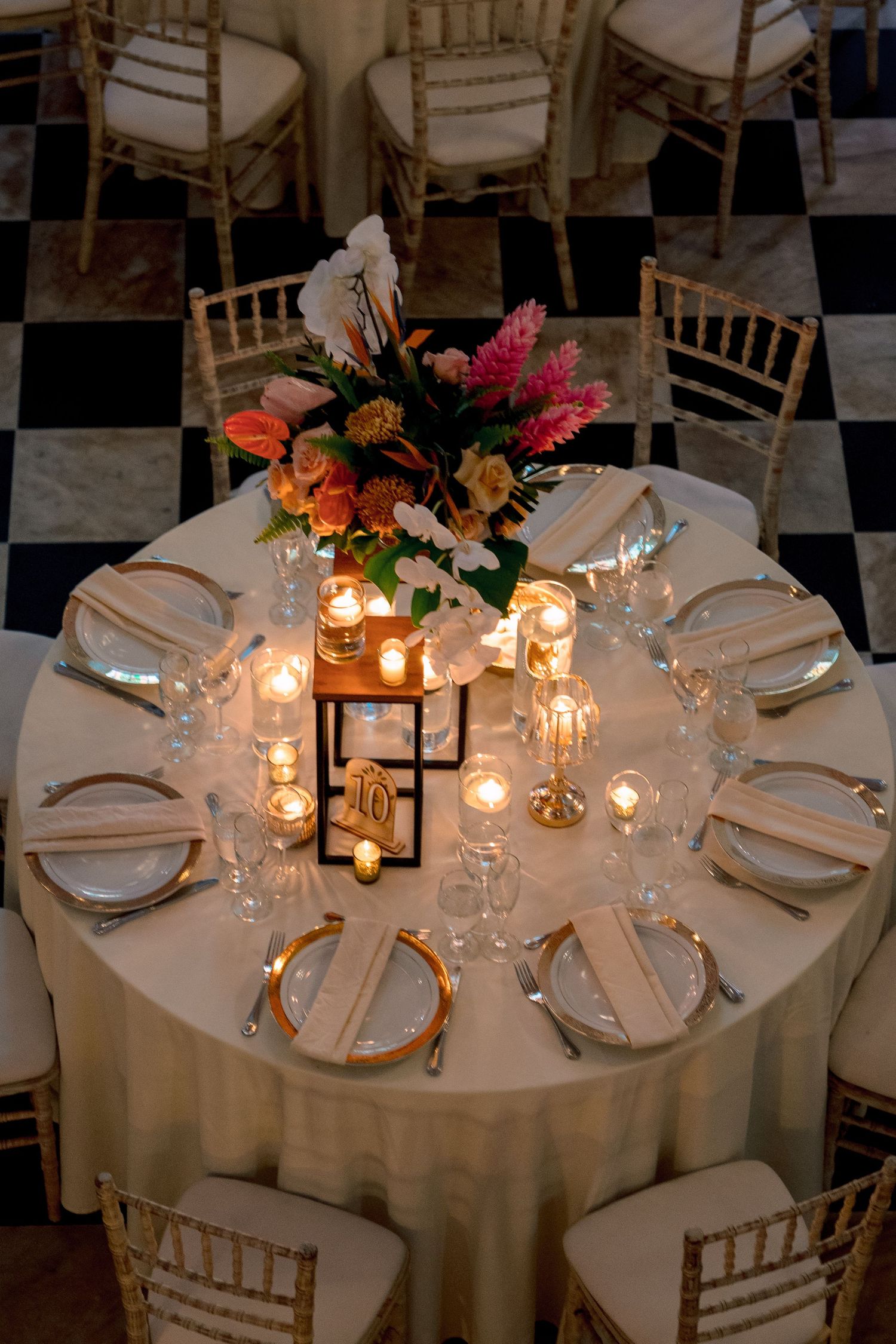 Elegant round wedding reception table with gold chargers, candles, and vibrant tropical floral centerpiece.