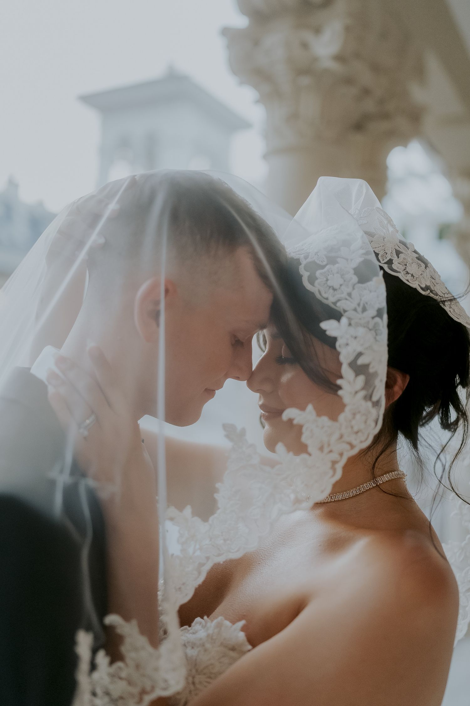 Bride and groom touching foreheads romantically under lace bridal veil in soft dreamy outdoor setting.