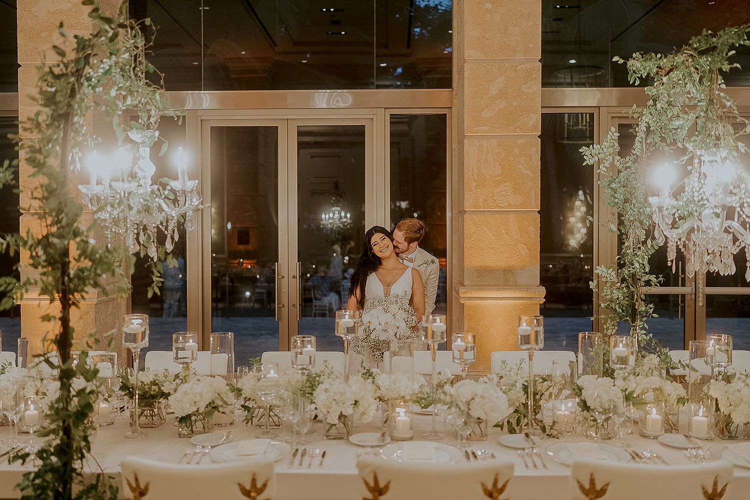 Couple embraces at elegant candlelit wedding reception with lush greenery and white floral centerpieces.