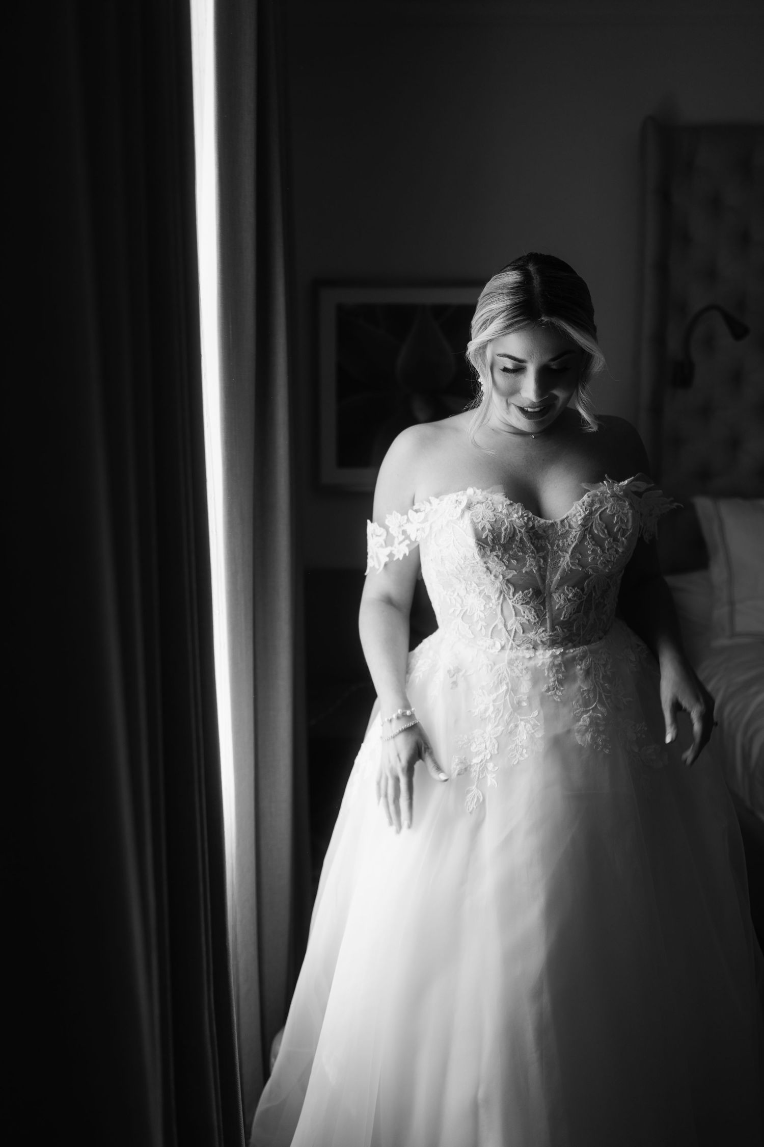 Bride in off-shoulder lace ballgown wedding dress poses by window in elegant black and white portrait photo.