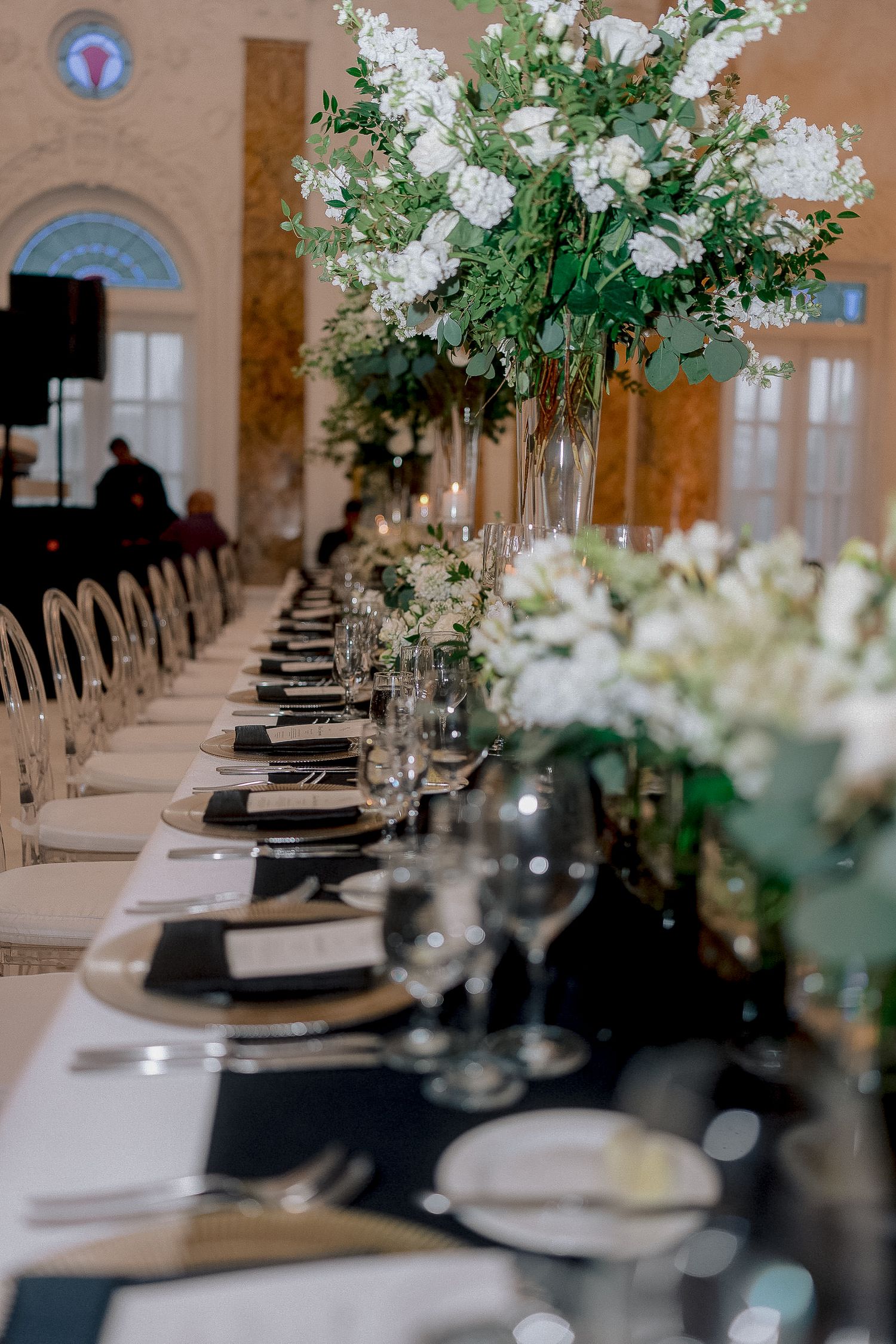 Long wedding reception table with tall white floral centerpieces, black linens, gold charger plates, and crystal glasses.