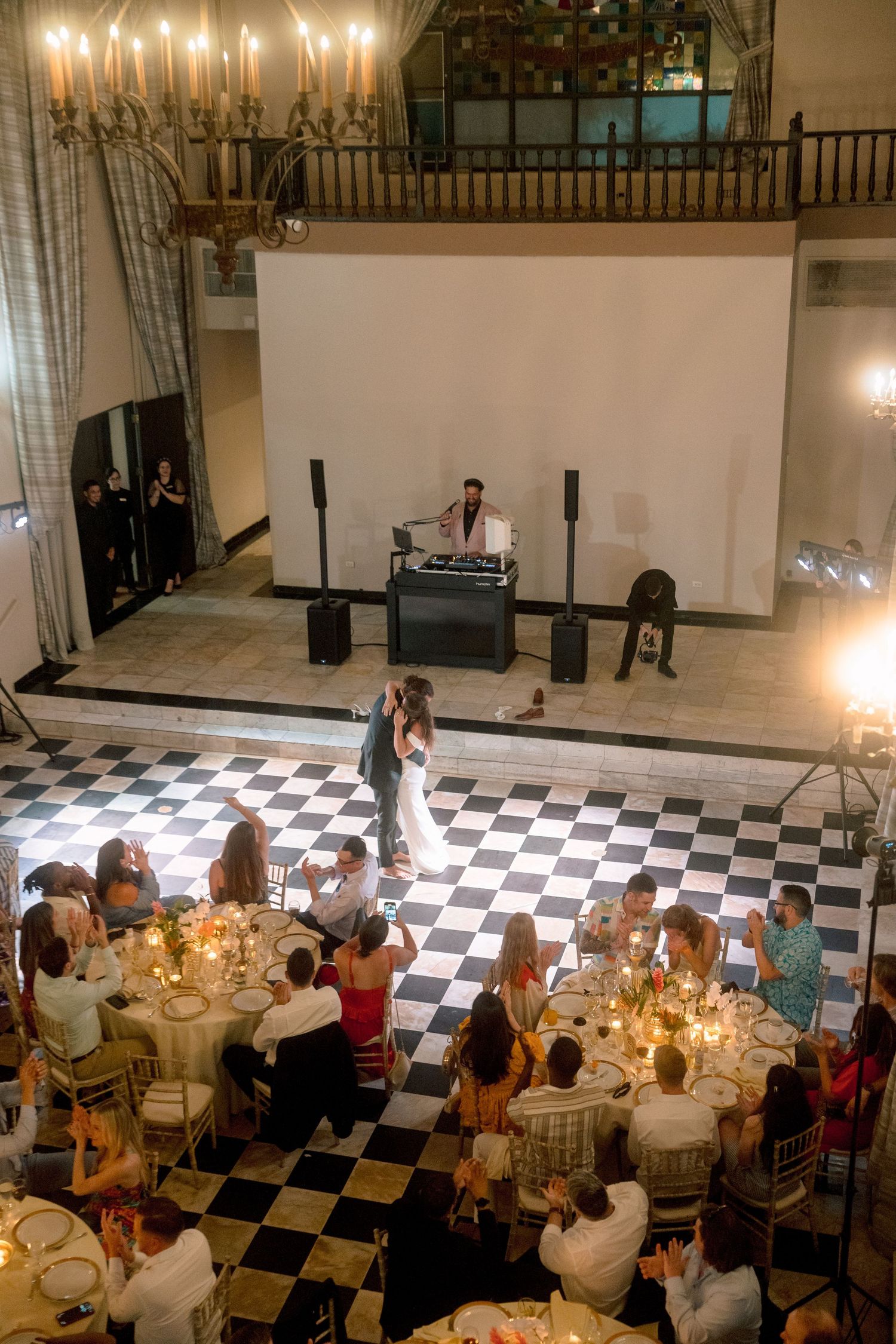 Aerial view of wedding reception with couple dancing on checkered floor and DJ performing on stage.