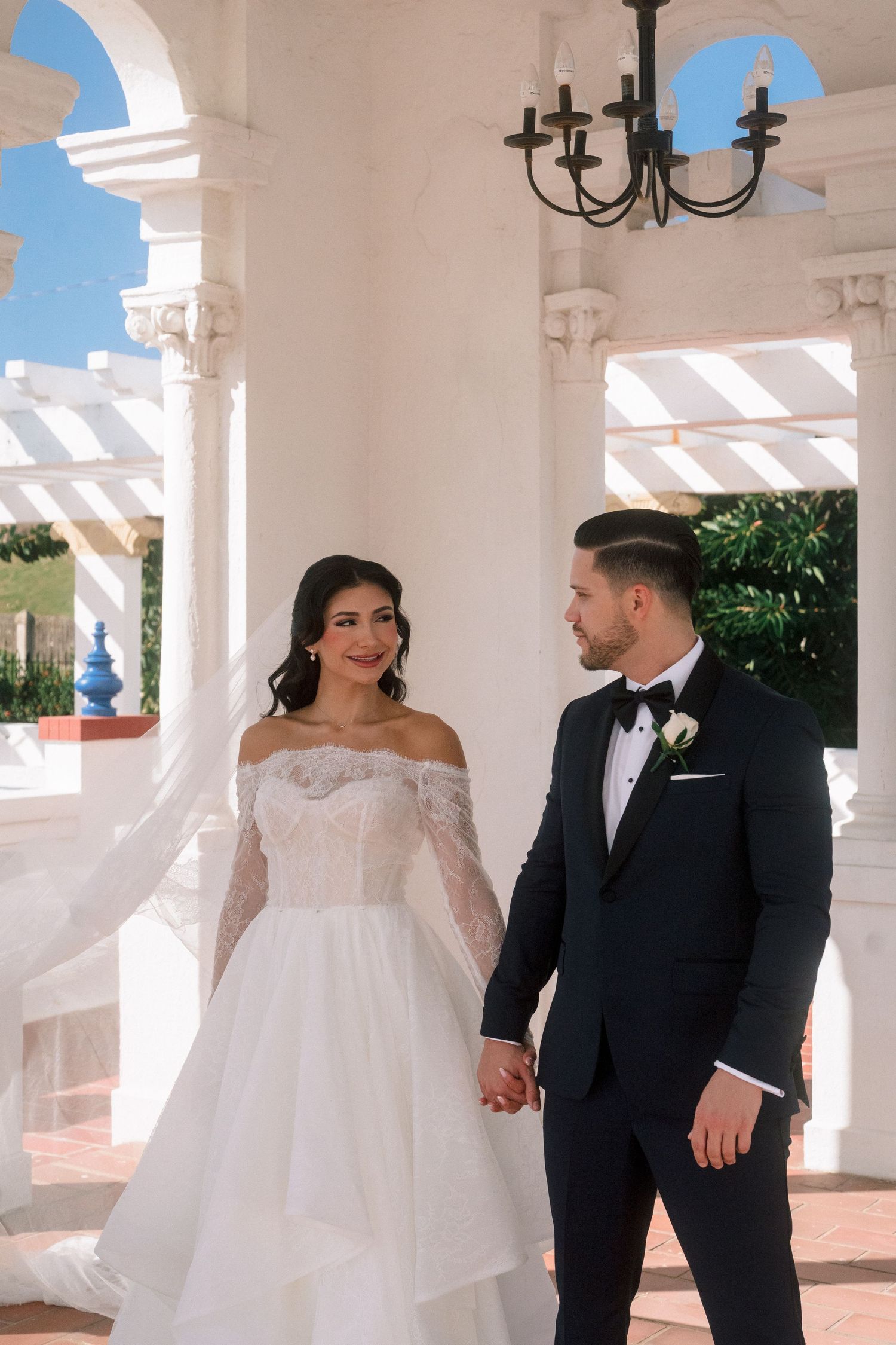 Bride in off-shoulder lace gown holds hands with groom in black tuxedo under white columns at elegant wedding venue.