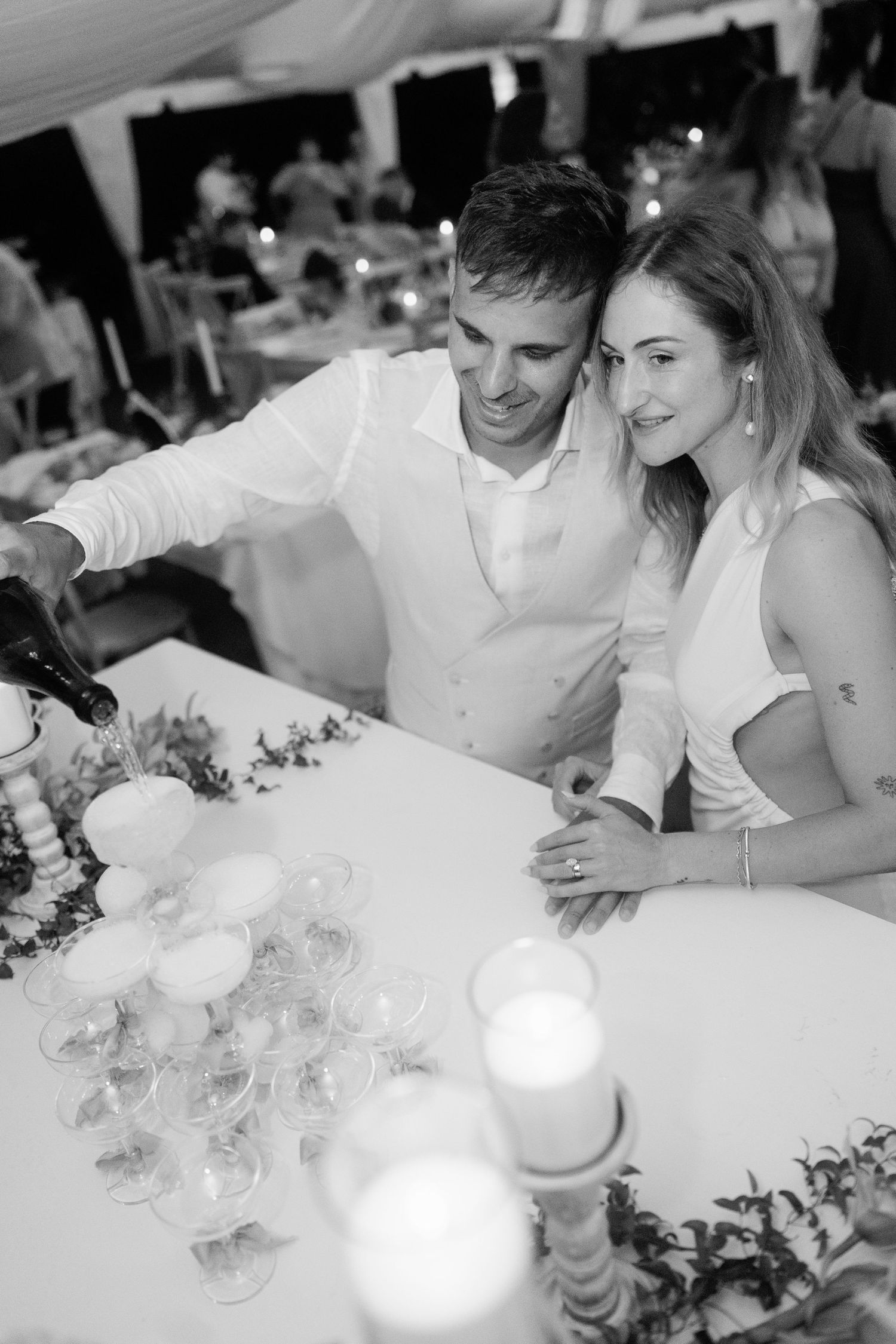 Black and white photo of newlyweds pouring champagne tower together at candlelit wedding reception.