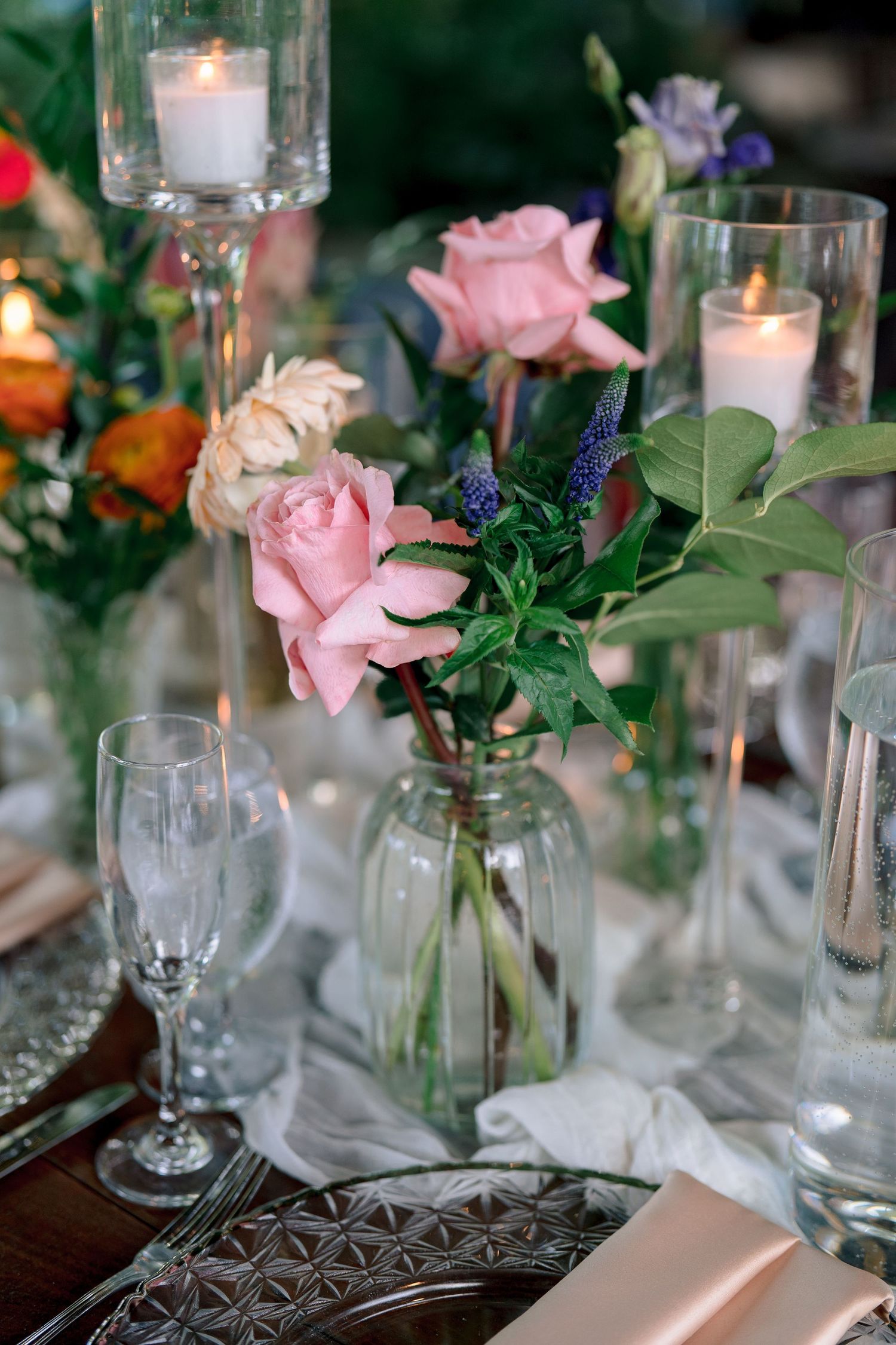 Elegant wedding table setting with pink roses in glass bud vases, candles, and crystal glassware.