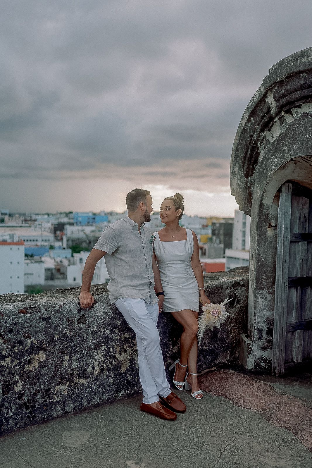 Couple in white attire share romantic moment on ancient stone fortress overlooking city.