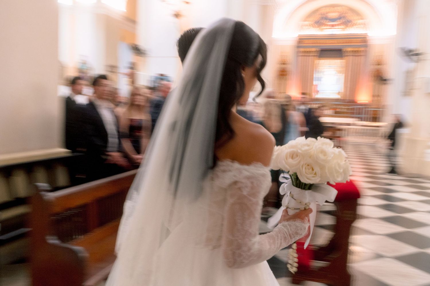 Bride in white lace gown and veil walks down church aisle holding white bouquet during wedding ceremony.