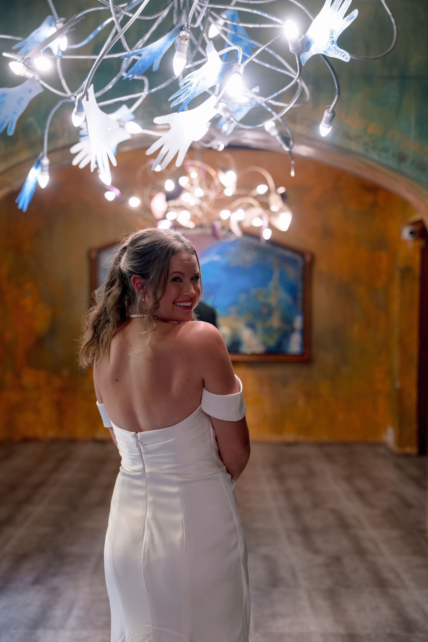 Smiling bride in white off-shoulder gown poses under glowing hand chandelier in elegant rustic venue.