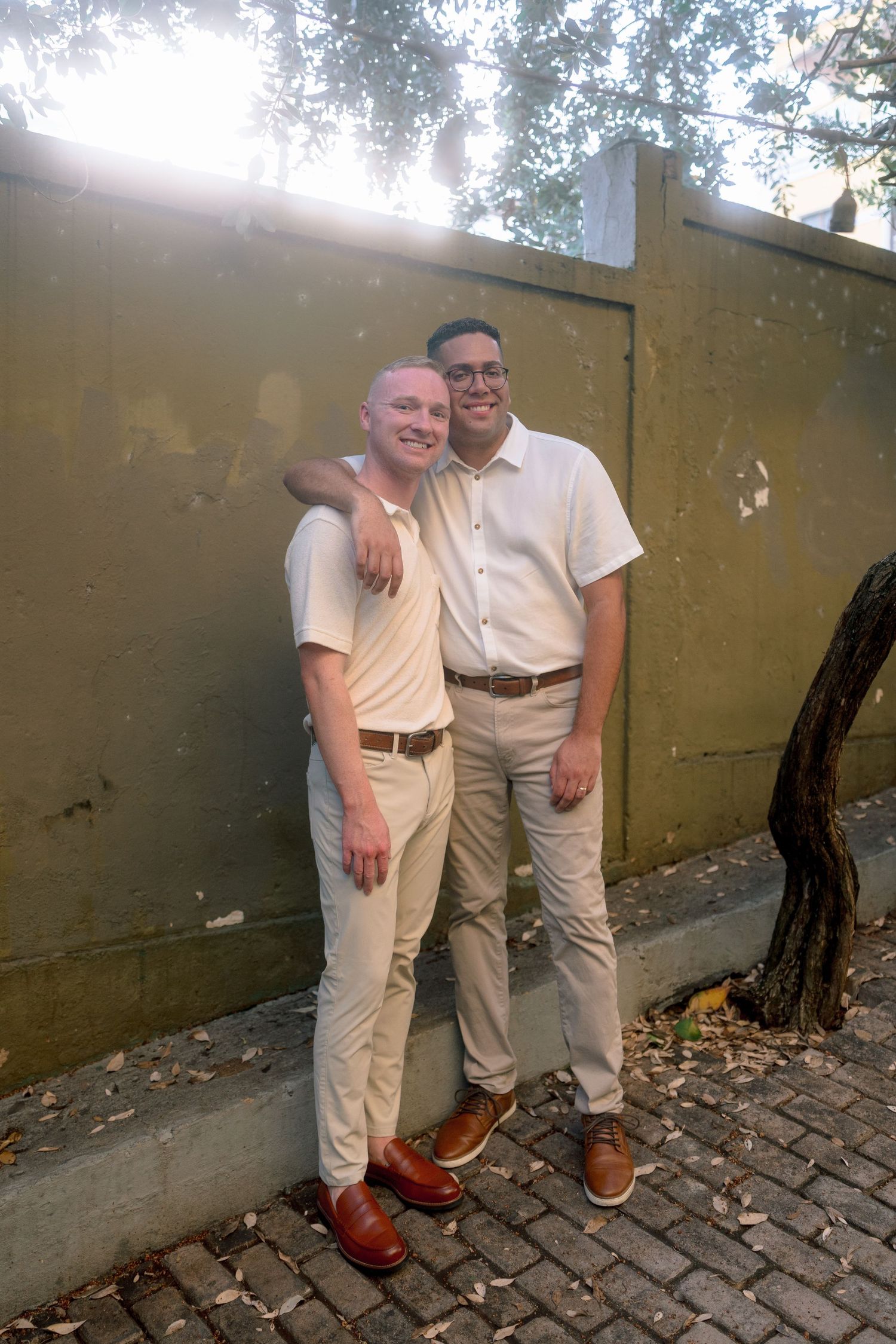 Two men in matching white shirts and khaki pants pose together outdoors against a green wall on a cobblestone path.