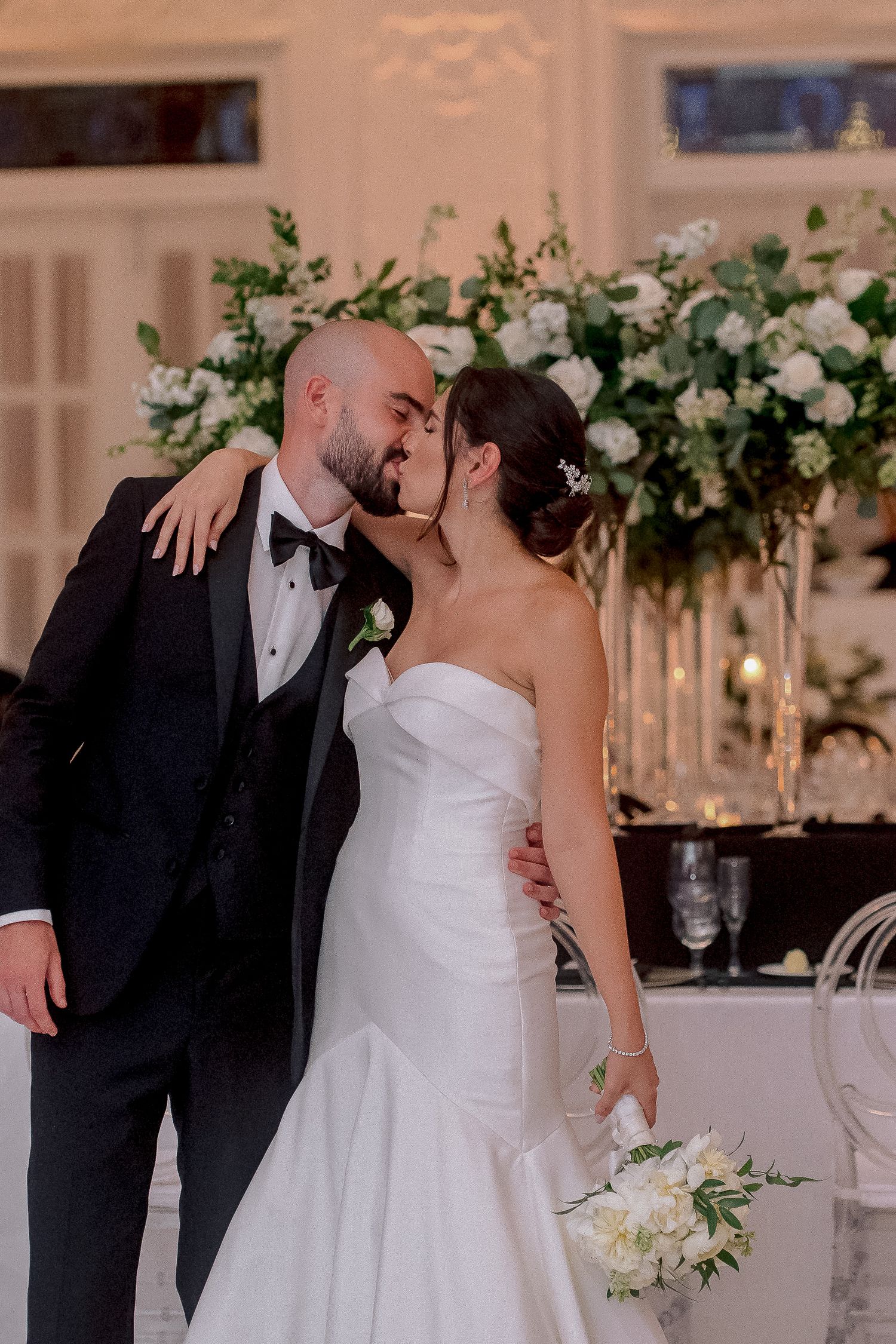 Bride and groom sharing a kiss at wedding reception, white floral arrangements, bride in strapless gown holding bouquet.
