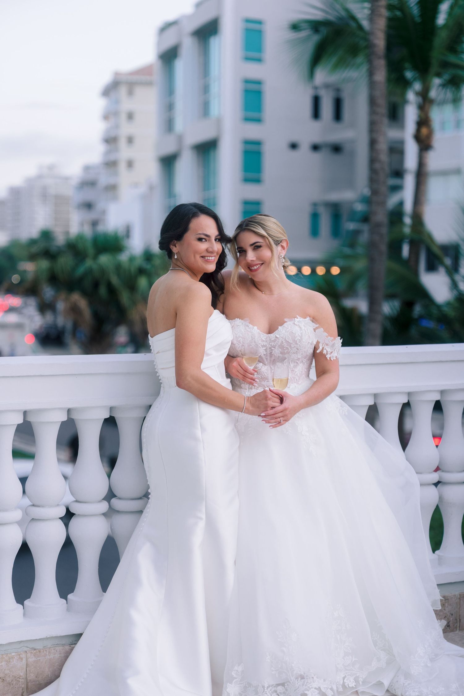 Two brides in white wedding gowns pose on a balcony with champagne, palm trees and buildings in the background.
