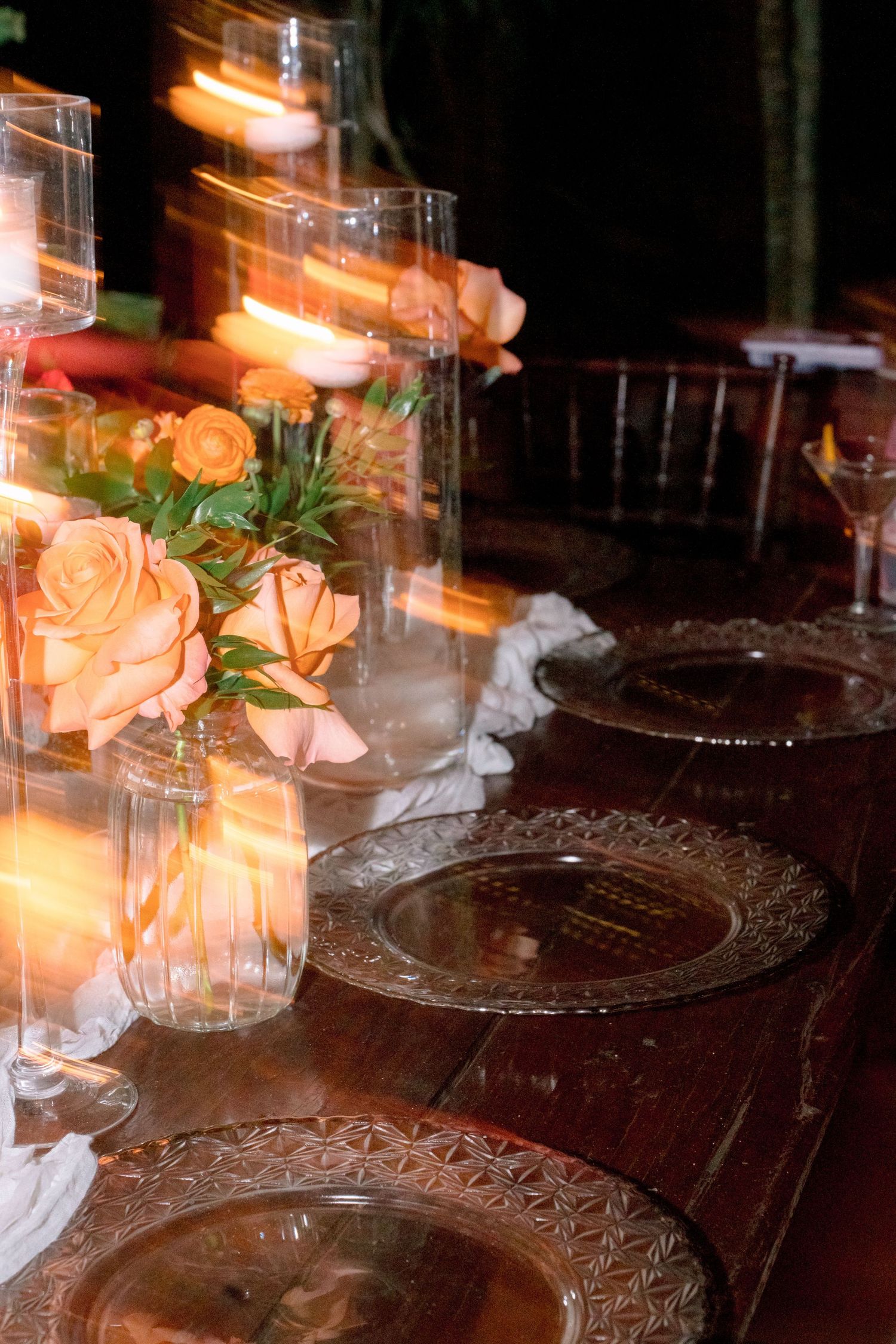 Elegant wedding table setting with peach roses, crystal charger plates, and glowing candles in tall glass holders.