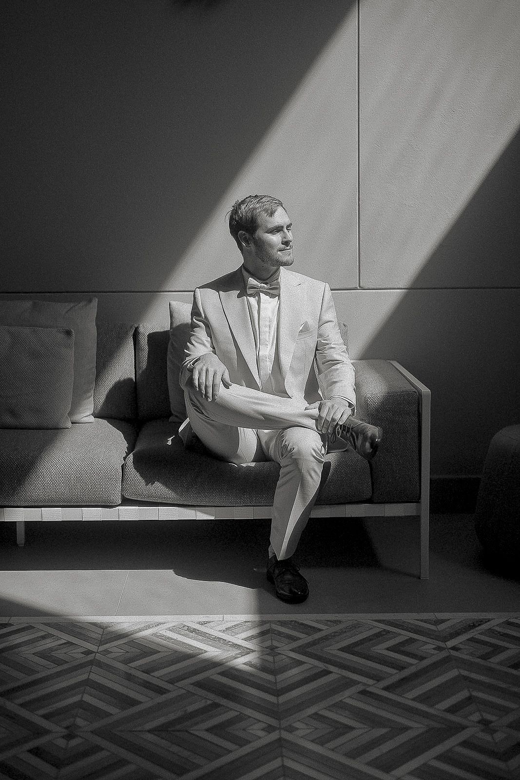 Elegant groom in a light suit sitting on a modern sofa in dramatic black and white lighting.