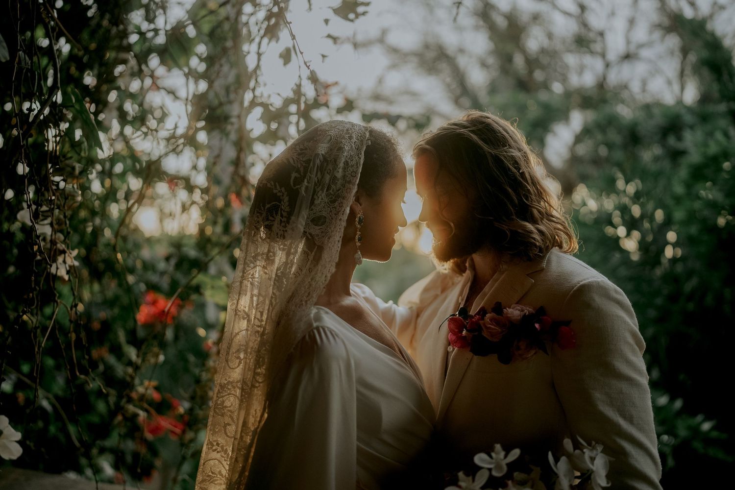 Two brides face each other tenderly in a sunlit garden surrounded by blooming flowers and lush greenery.