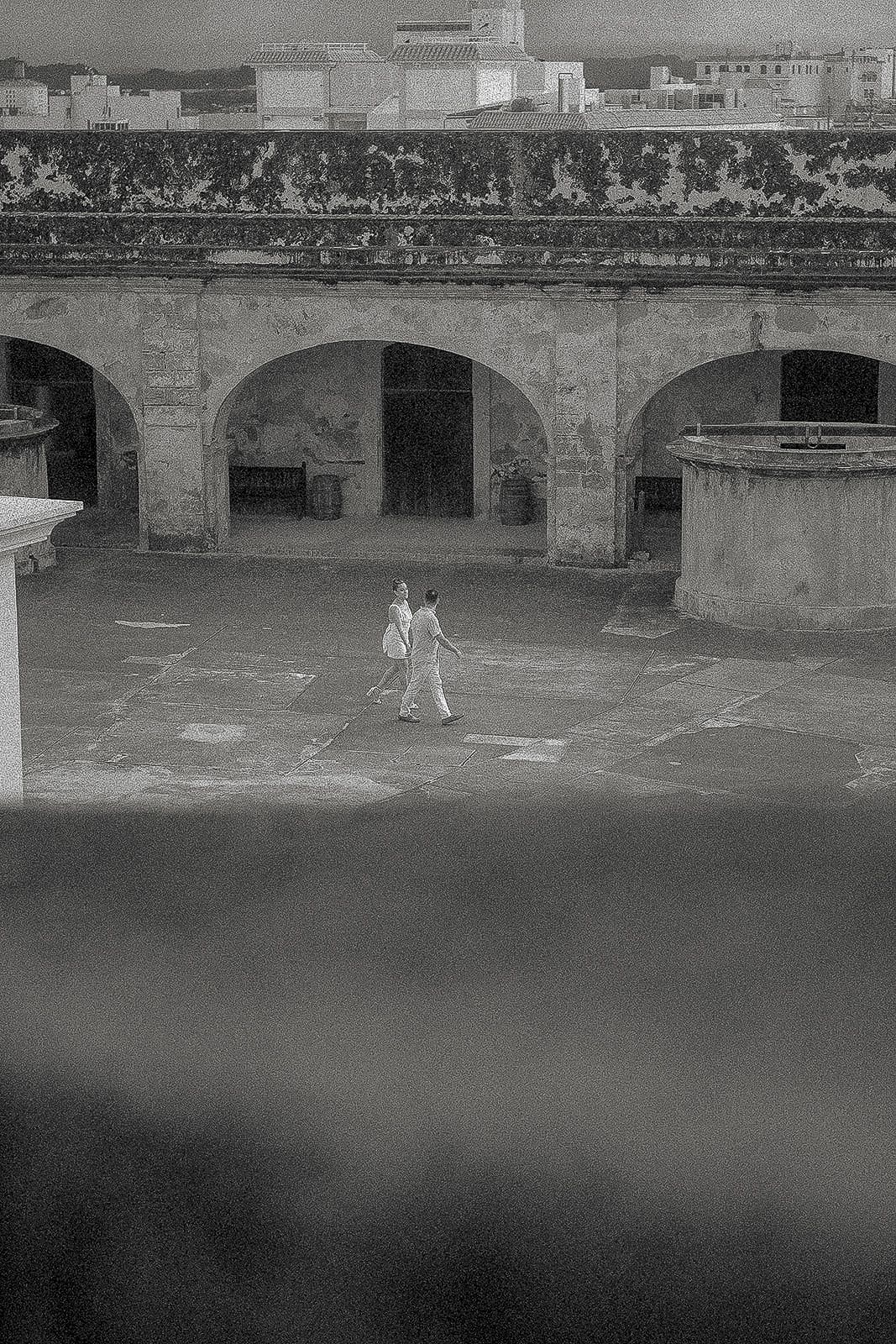 Black and white photo of two people walking across a historic courtyard with stone arches in an old colonial building.
