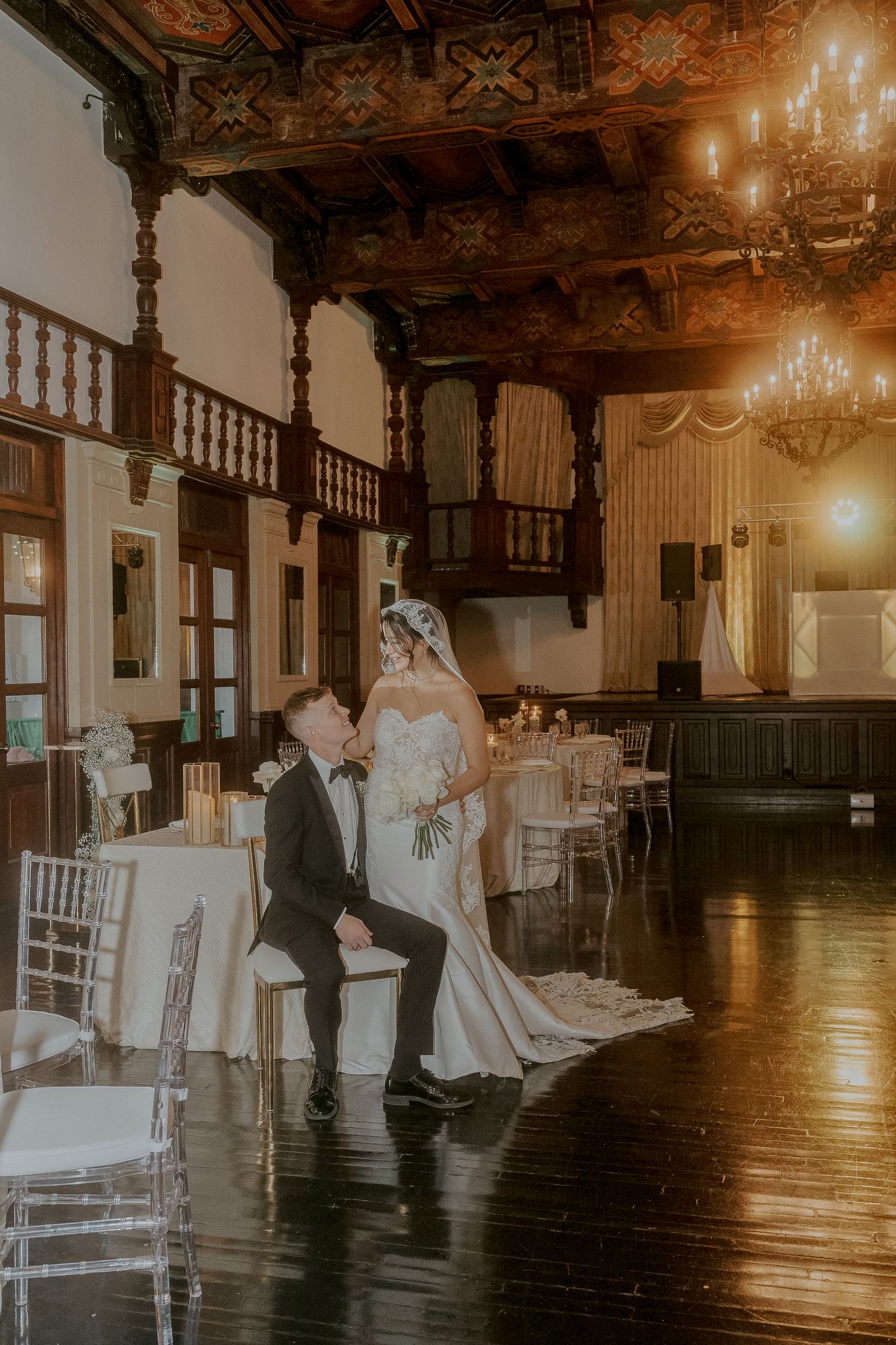 Bride and groom pose elegantly in a grand ballroom with chandeliers.